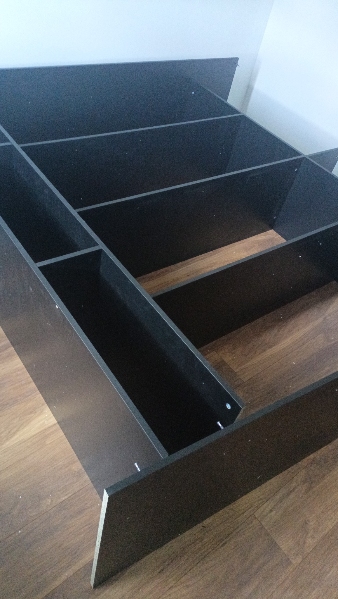 Częściowo zmontowana, czarna szafa regałowa na podłodze z paneli, widoczne śruby montażowe, wnętrze półek w kolorze drewna. Ujęcie z dołu, pod kątem.