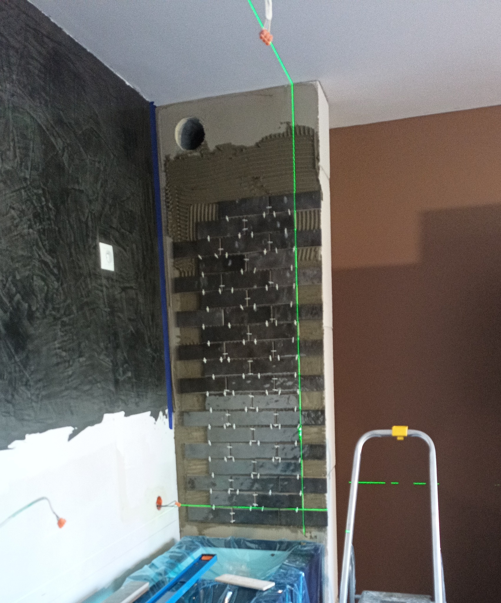 Wnętrze w trakcie wykończenia: fragment ściany z ułożonymi ciemnymi płytkami, krzyżyki dystansowe, poziomica laserowa, widoczna drabina i zabezpieczone elementy pomieszczenia.