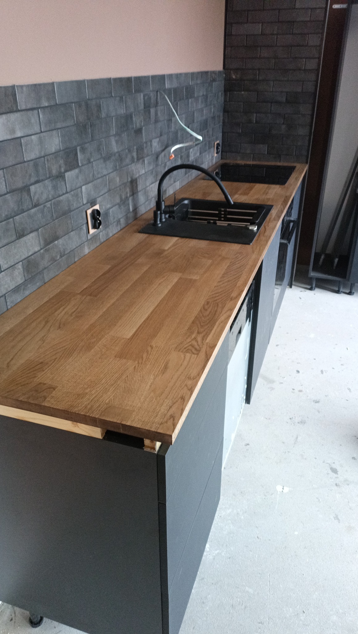 Częściowy widok blatu z drewna i czarnego zlewu w nowoczesnej kuchni z szarą cegłą na ścianie. Widoczne niedokończone podłączenia elektryczne.