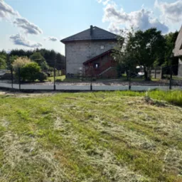 Nowoczesne ogrodzenie panelowe z podmur&oacute;wką, otaczające posesję z domami w tle. Widoczny fragment trawnika i zaparkowany samoch&oacute;d dostawczy.