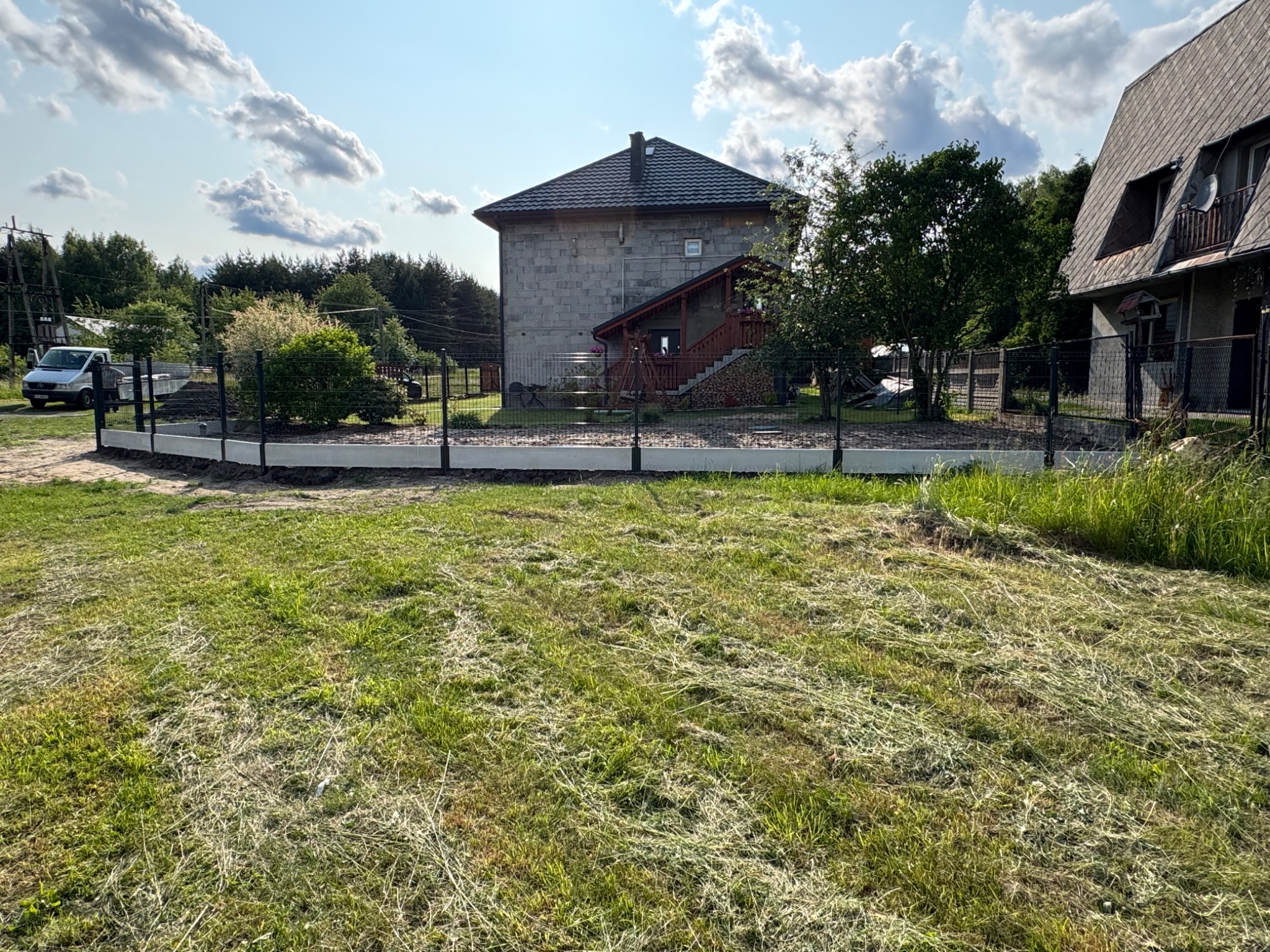 Nowoczesne ogrodzenie panelowe z podmurówką, otaczające posesję z domami w tle. Widoczny fragment trawnika i zaparkowany samochód dostawczy.