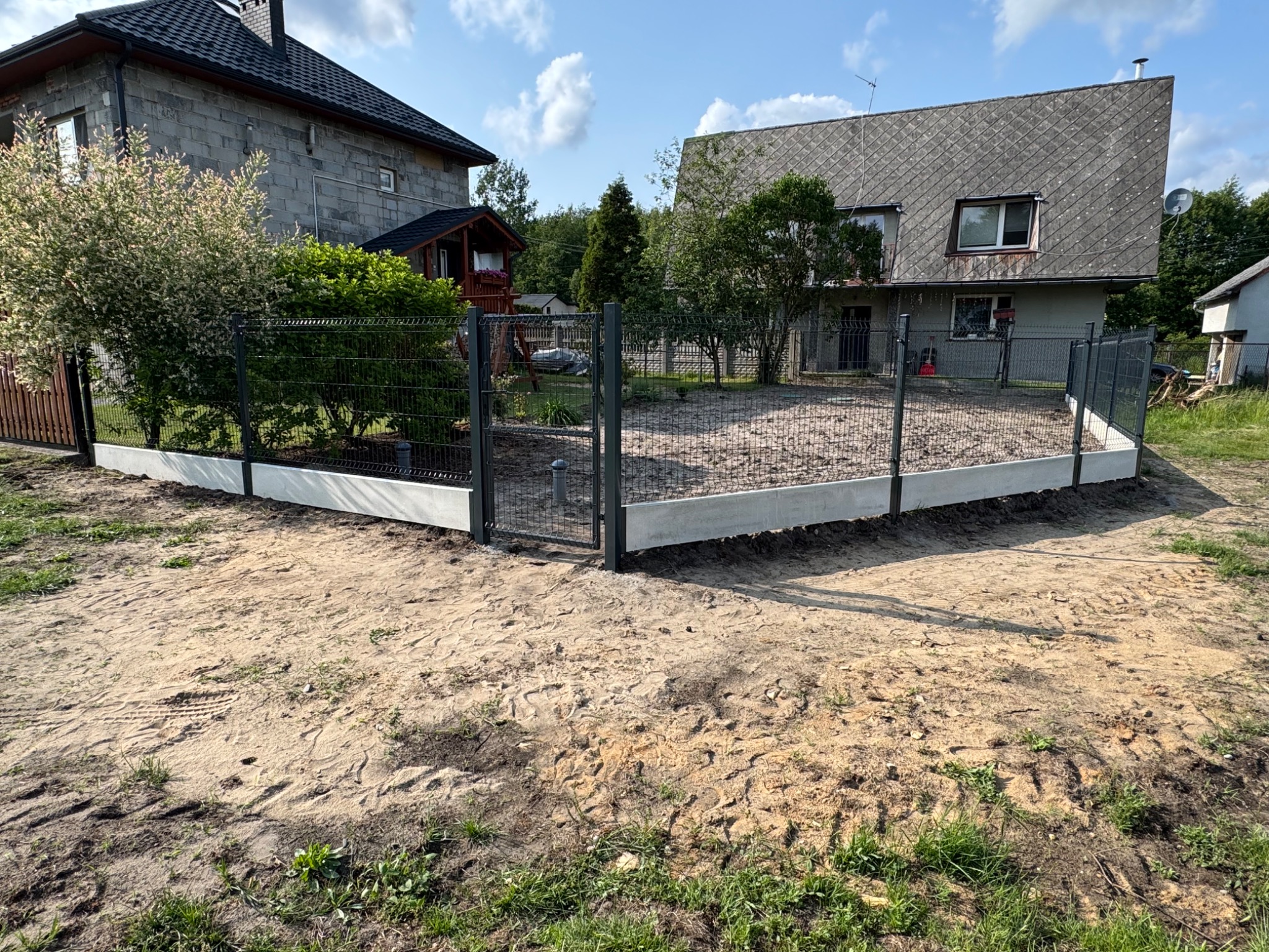 Nowoczesne ogrodzenie panelowe w kolorze grafitowym, osadzone na betonowej podmurówce, otaczające posesję z dwoma domami w tle, w słoneczny dzień.