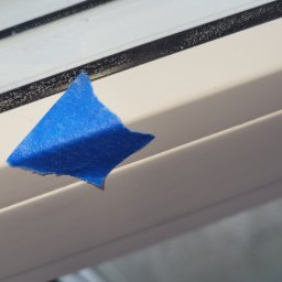 Michał Stępiński - Detal okna z niebieską taśmą malarską, widoczne zabrudzenia na ramie okiennej. Przygotowanie do malowania lub zabezpieczenie przed zabrudzeniem. Okno od wewnątrz.