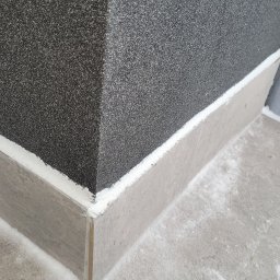 Michał Stępiński - Szaro-czarna elewacja budynku z dekoracyjnym cokołem z płytek, wypełniona białą fugą. Widoczny narożnik i fragment posadzki.