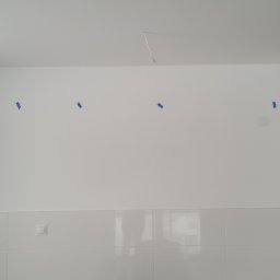 Michał Stępiński - Biała ściana z płytkami i fragmentem wystającej instalacji elektrycznej z sufitu, zaznaczone miejsca taśmą malarską. Widoczne gniazdka.