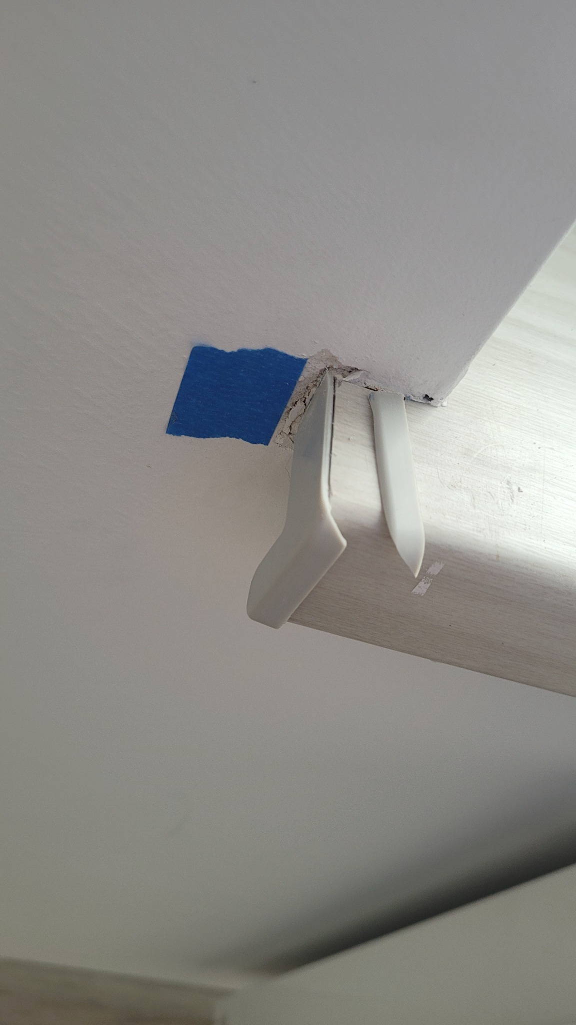 Fragment sufitu z niebieską taśmą malarską, uszkodzonym narożnikiem i odklejonym elementem wykończeniowym. Widoczne szczeliny i niedociągnięcia.