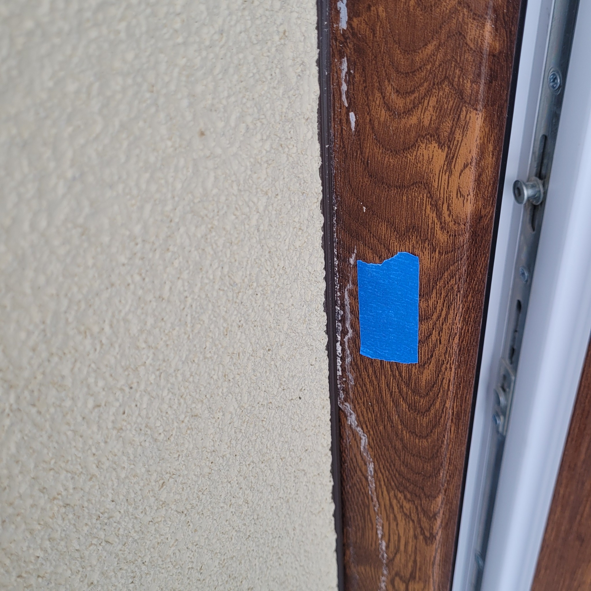 Fragment okna z brązową ramą imitującą drewno, zabezpieczoną niebieską taśmą malarską, przylegającą do ściany o jasnej, ziarnistej strukturze.