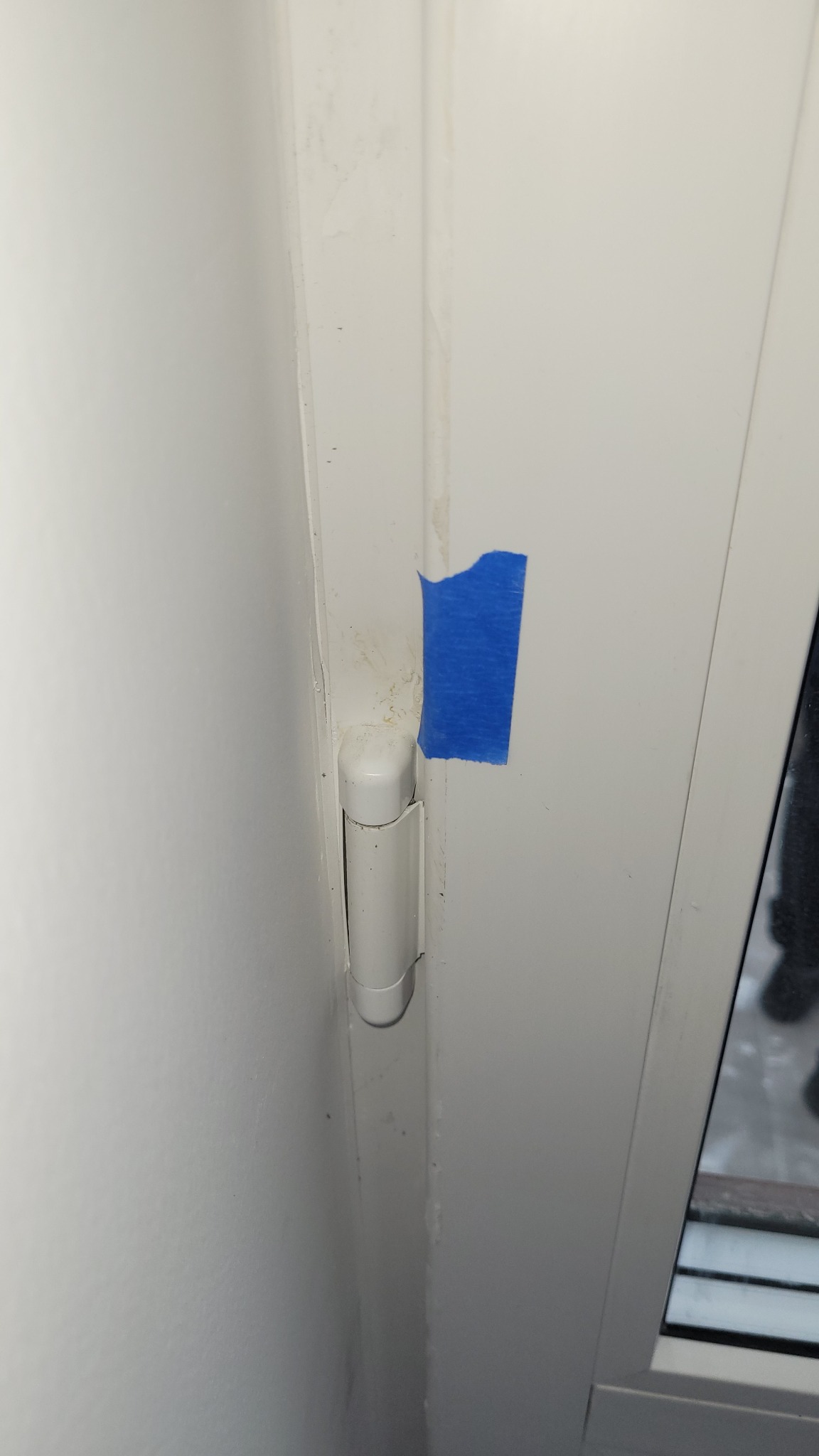 Białe zawiasy okienne zabezpieczone niebieską taśmą malarską, widoczne ślady po wcześniejszych pracach. Detal okna po renowacji.