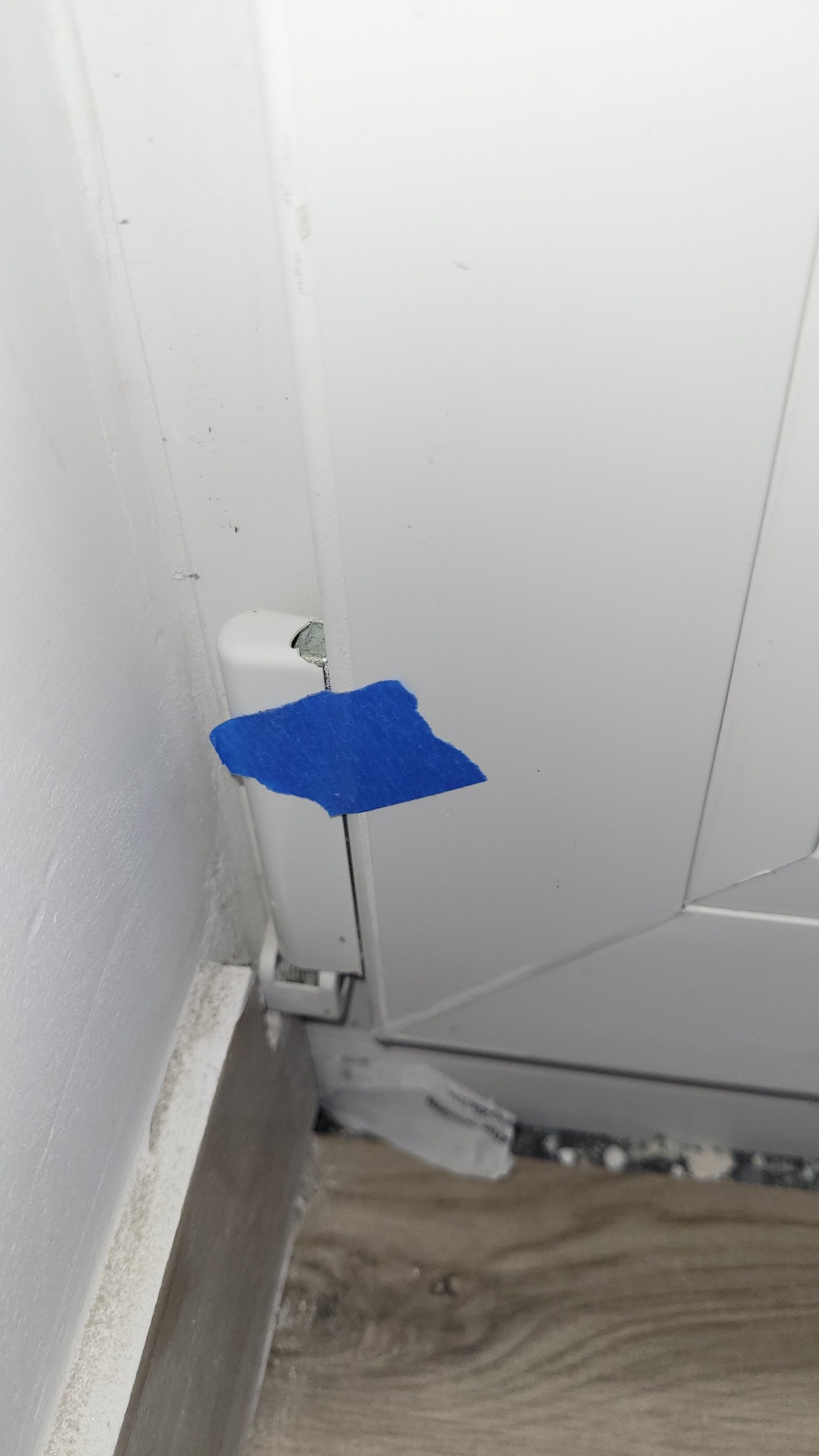 Szczegółowe ujęcie uszkodzonego okna PCV z niebieską taśmą zabezpieczającą, ukazujące defekt i niedoróbki wykończeniowe przy podłodze.