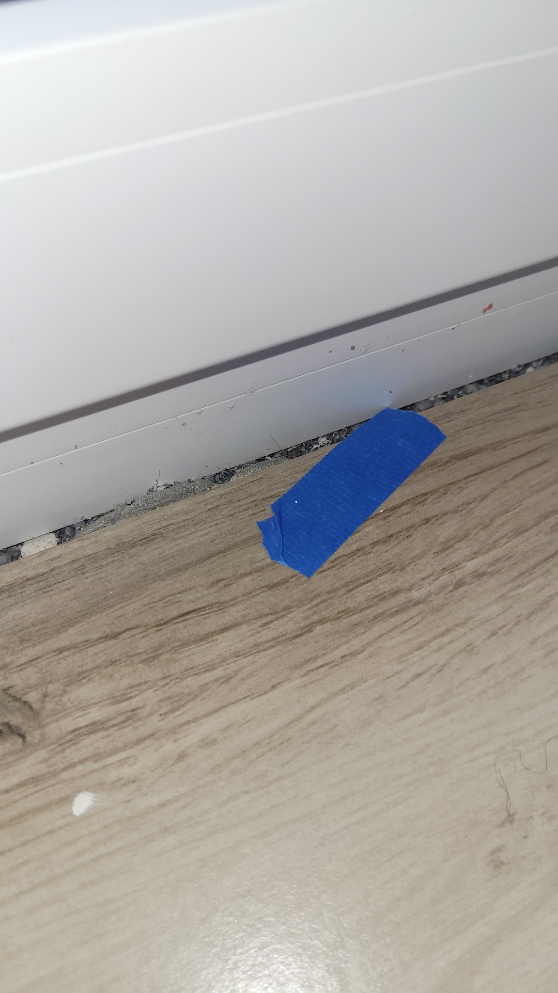 Niedokładne wykończenie podłogi przy listwie przypodłogowej, zabezpieczone niebieską taśmą malarską na panelach podłogowych.
