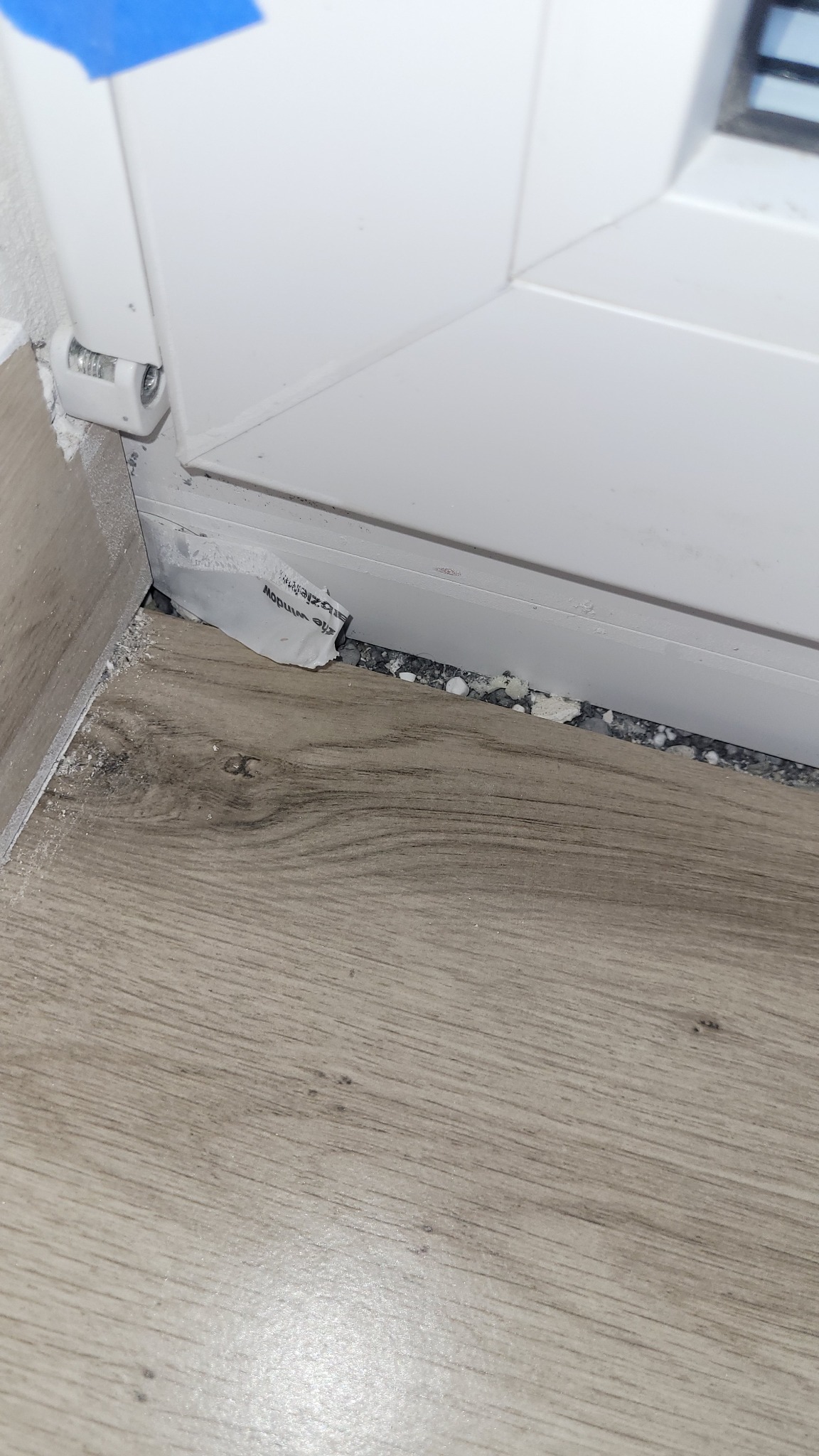 Szczegółowe ujęcie nieszczelnego okna z widoczną pianką montażową i fragmentem etykiety. Podłoga laminowana w neutralnym odcieniu drewna.