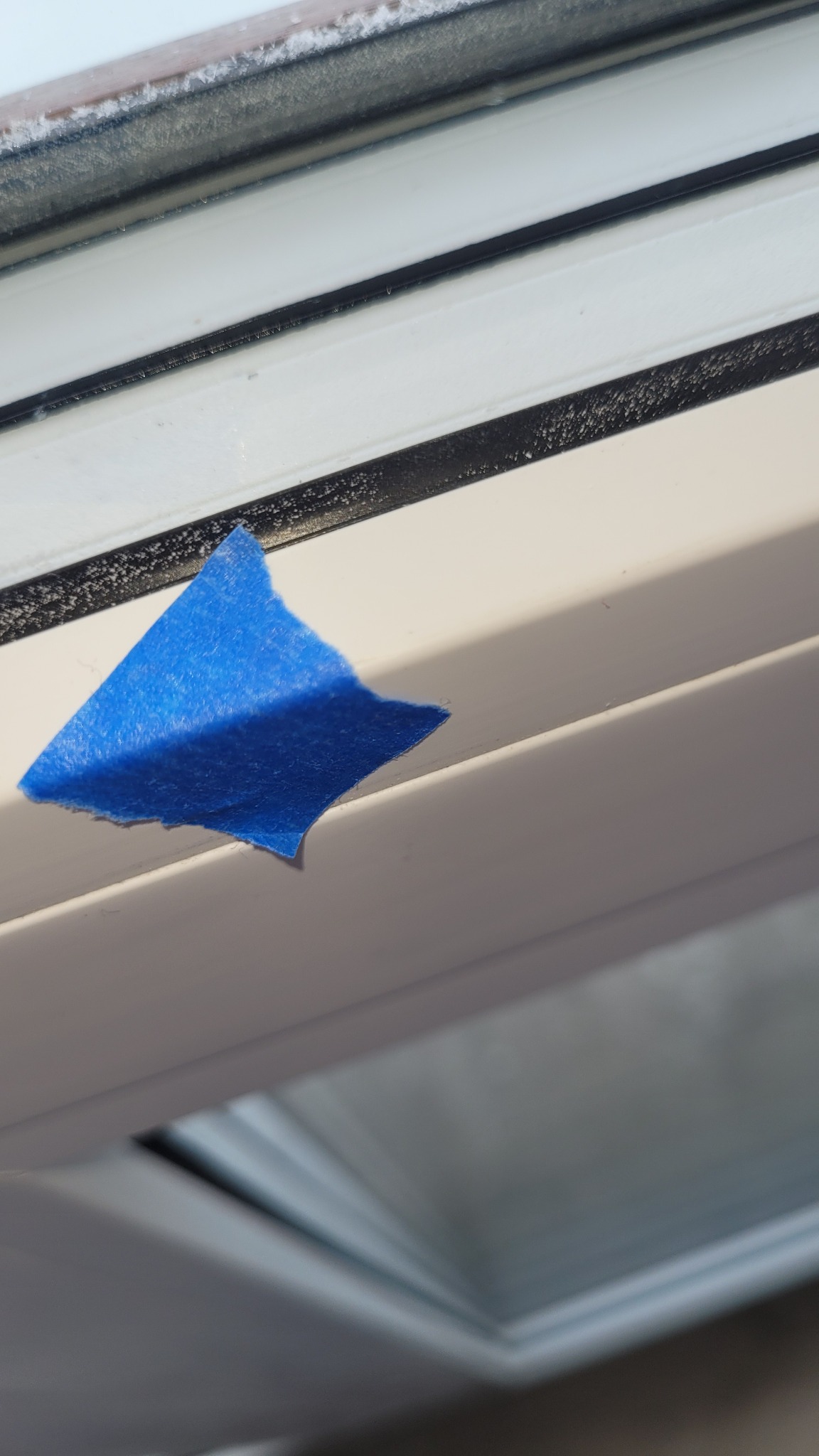 Detal okna z niebieską taśmą malarską, widoczne zabrudzenia na ramie okiennej. Przygotowanie do malowania lub zabezpieczenie przed zabrudzeniem. Okno od wewnątrz.