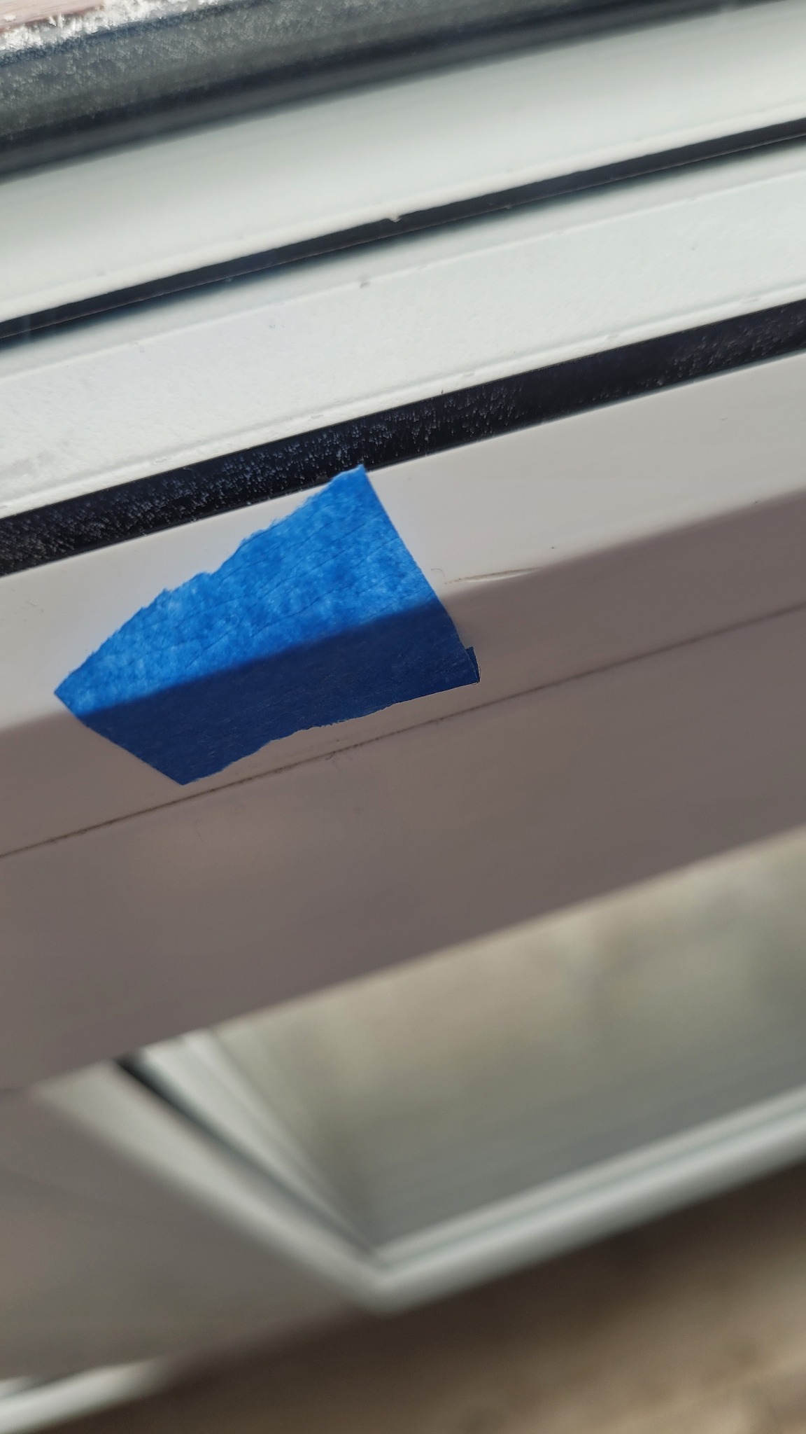 Fragment białej ramy okiennej z przyklejoną niebieską taśmą malarską, zabezpieczającą przed zabrudzeniem podczas prac wykończeniowych.
