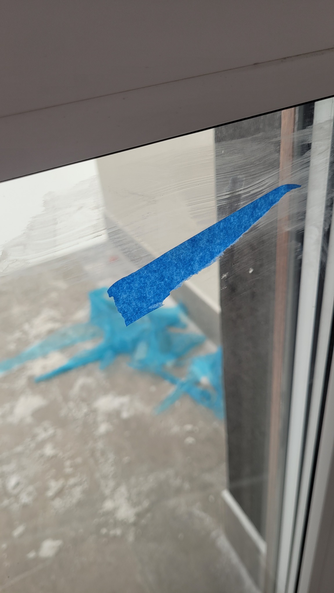 Okno zabezpieczone niebieską taśmą malarską, widoczne ślady po farbie i niebieska folia ochronna na podłodze. Przygotowanie do prac malarskich.