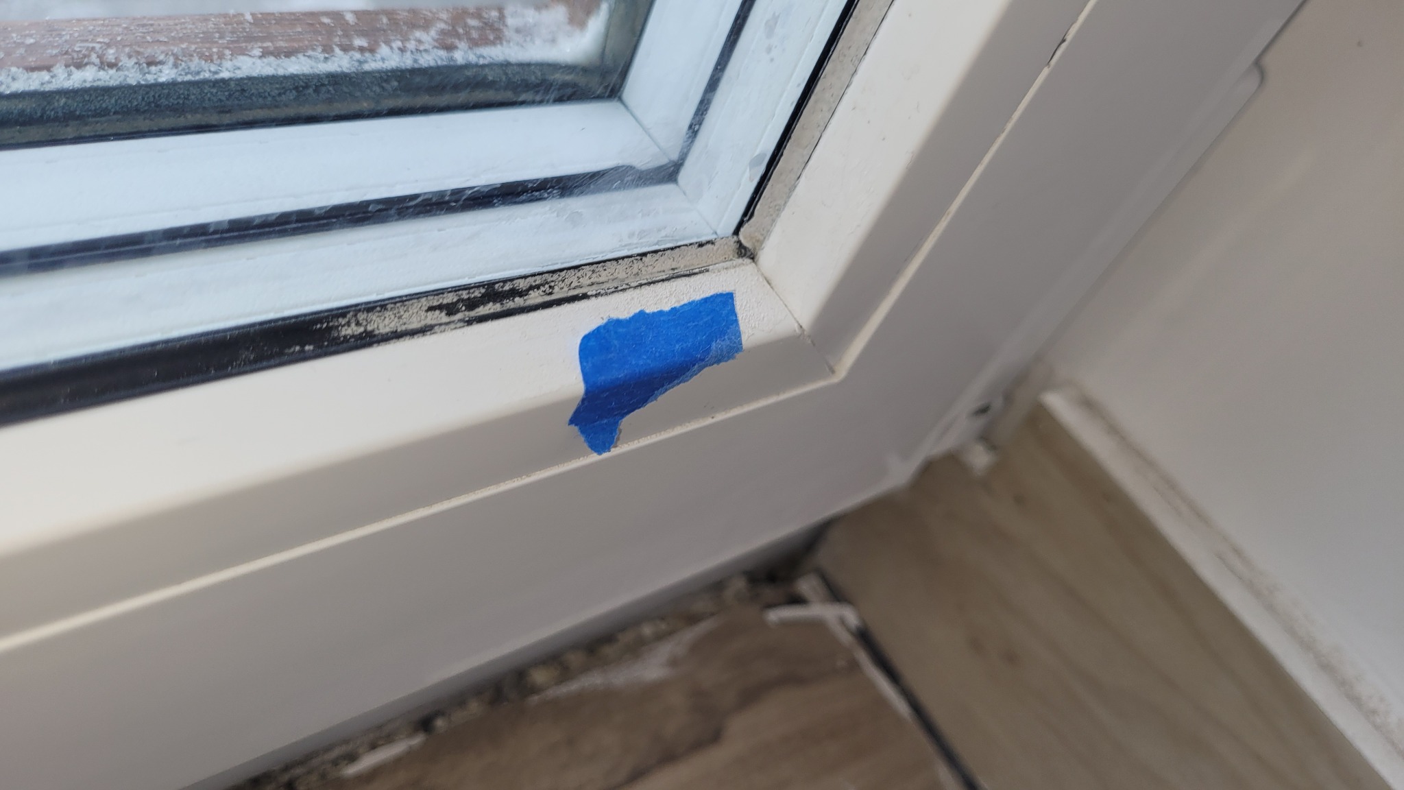 Szczegółowe ujęcie rogu okna PCV z niebieską taśmą malarską. Widoczne zabrudzenia na ramie okiennej i silikonie. Fragment drewnopodobnej podłogi w tle.