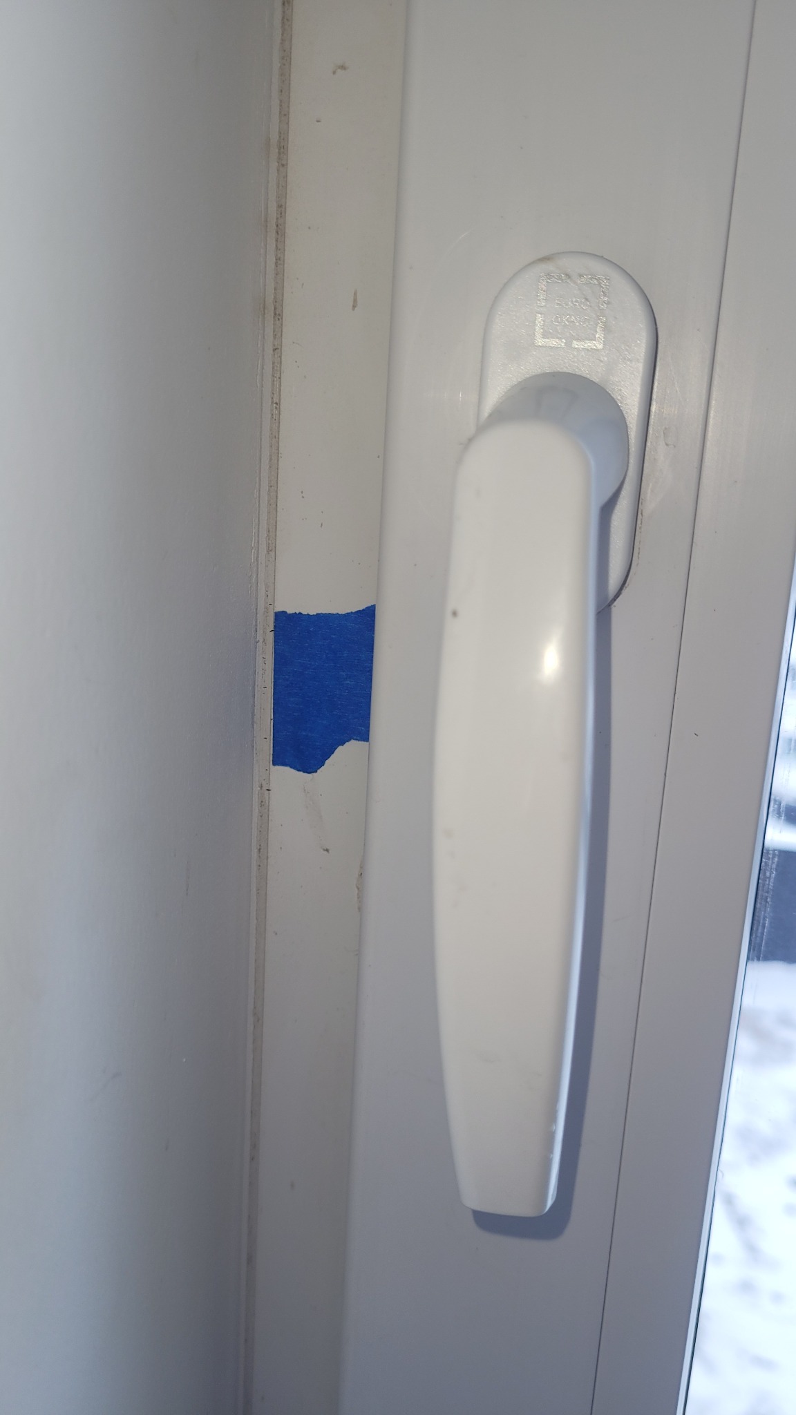 Białe okno PCV z klamką i fragmentem niebieskiej taśmy malarskiej na ramie, widoczne ślady zabrudzeń i uszkodzeń powłoki lakierniczej. Zbliżenie.