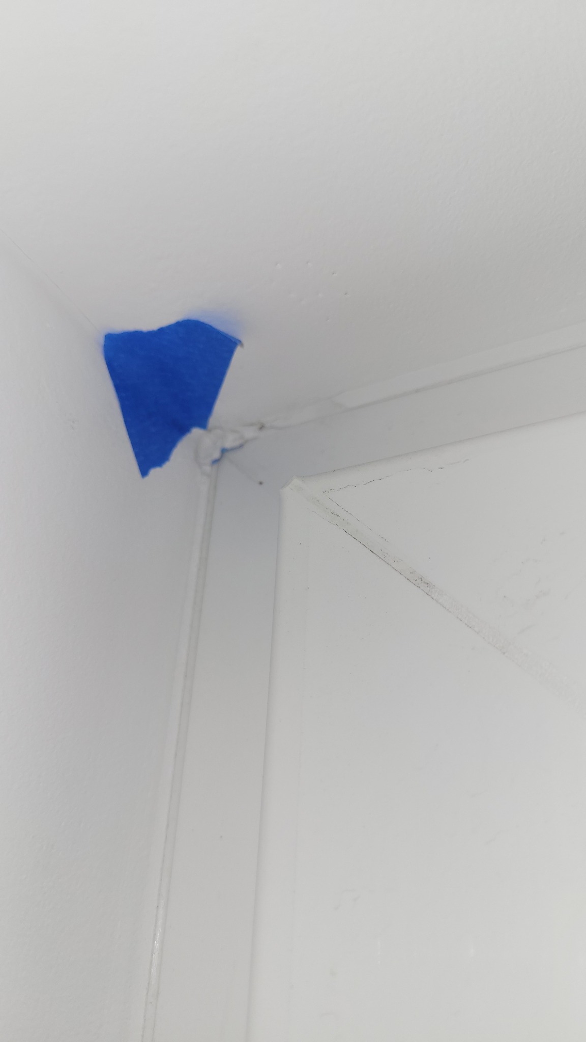 Biała ściana i sufit z niebieską taśmą malarską w rogu, zabezpieczającą przed zabrudzeniem farbą, obok widoczny fragment białych drzwi.