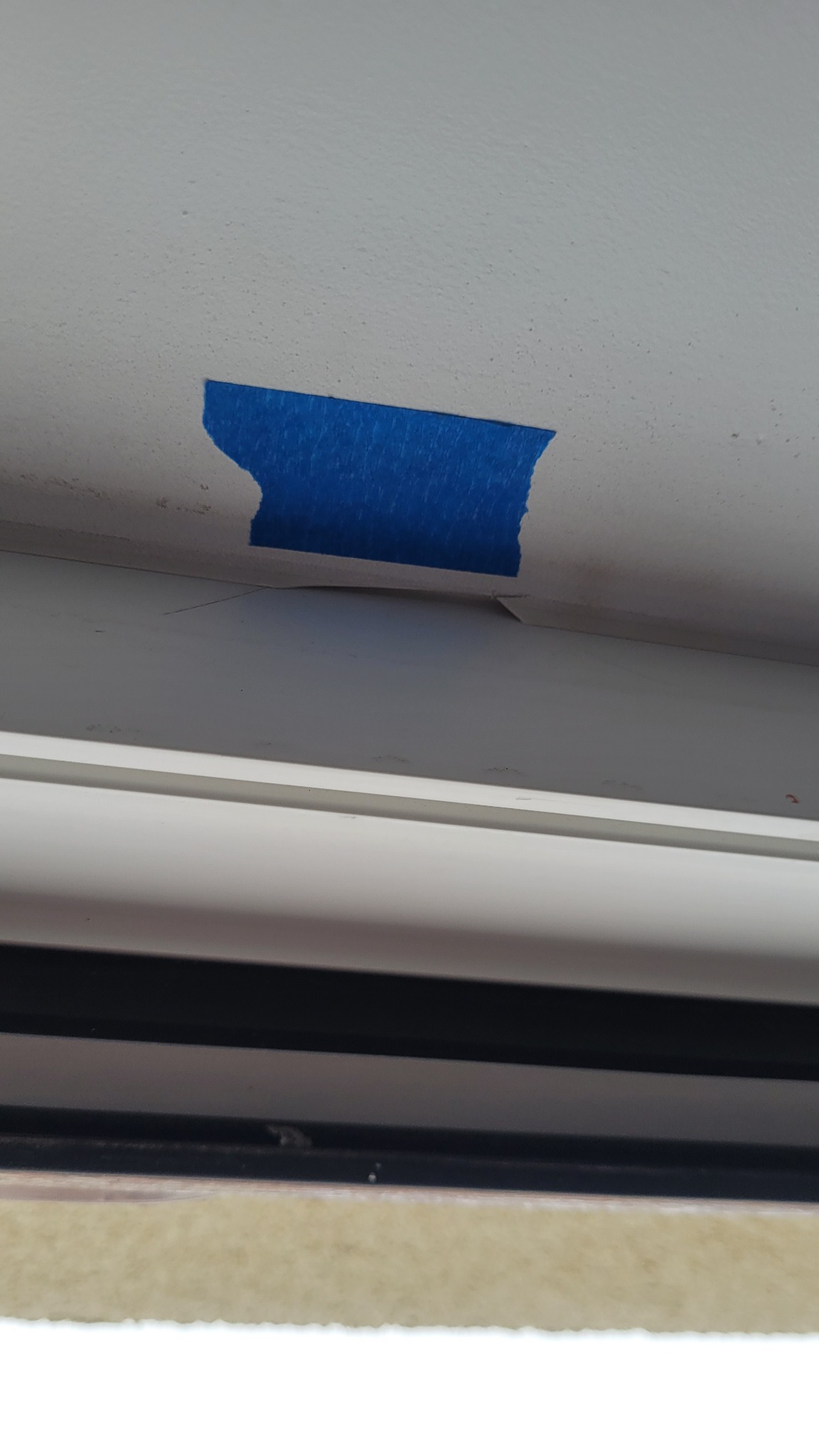 Niedokładne malowanie sufitu przy oknie, zabezpieczone niebieską taśmą malarską. Widoczne niedociągnięcia i nierówności.