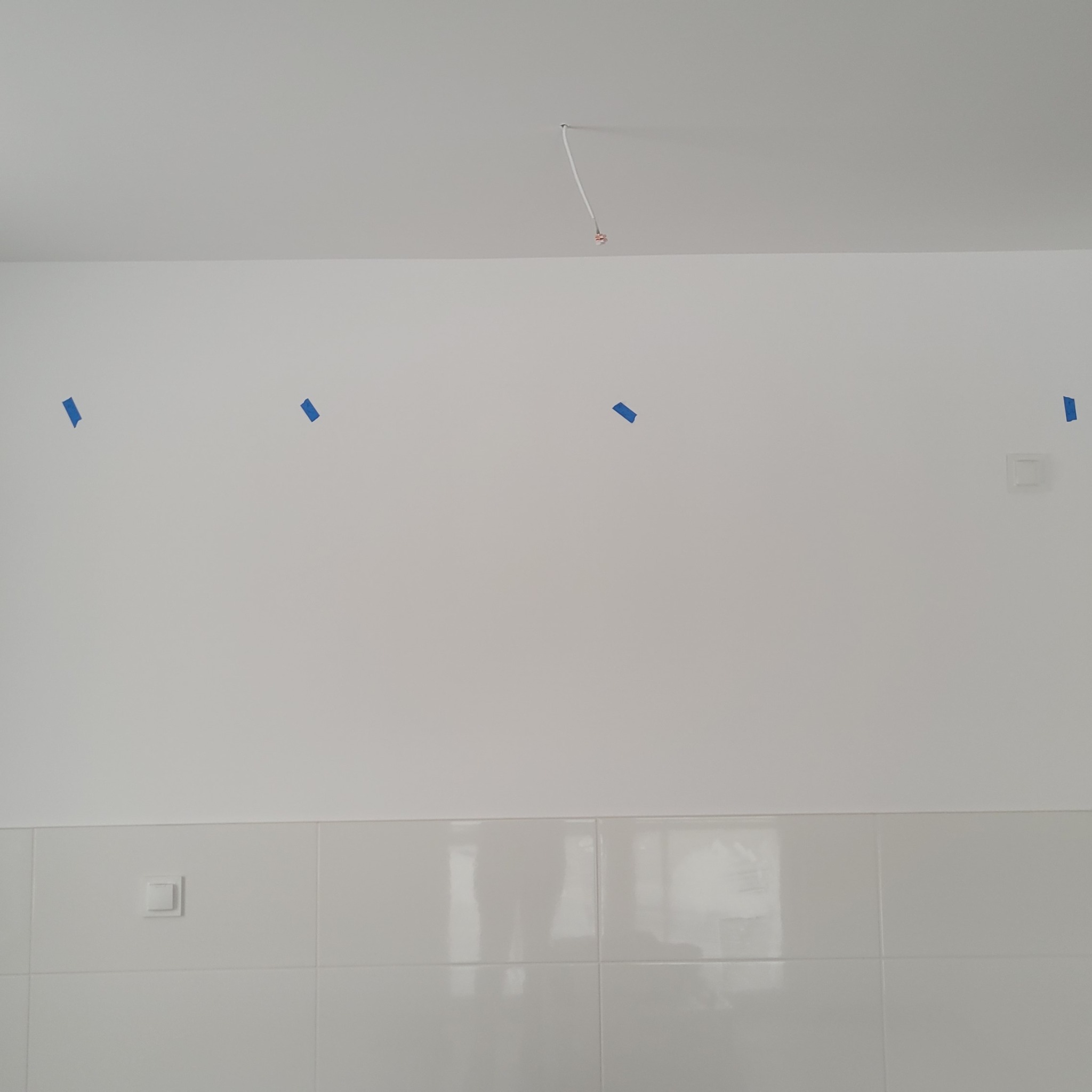 Biała ściana z płytkami i fragmentem wystającej instalacji elektrycznej z sufitu, zaznaczone miejsca taśmą malarską. Widoczne gniazdka.