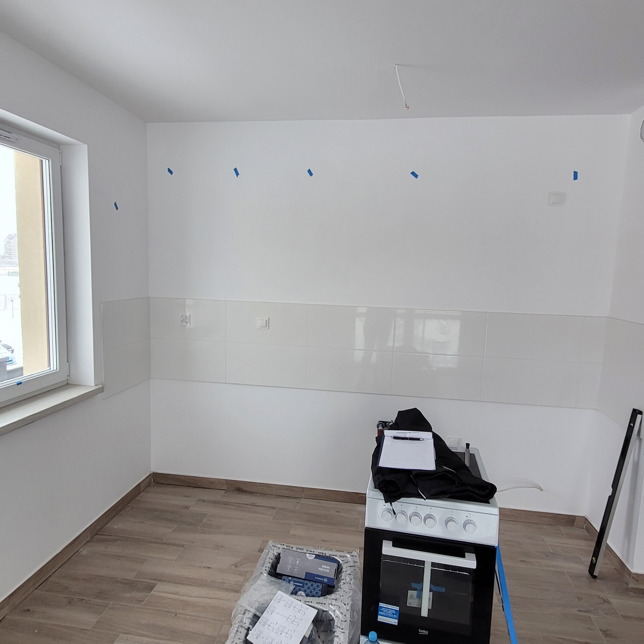 Pusta kuchnia z podłączoną płytą, świeżo położonymi płytkami i przygotowanymi punktami elektrycznymi oznaczonymi niebieską taśmą malarską.