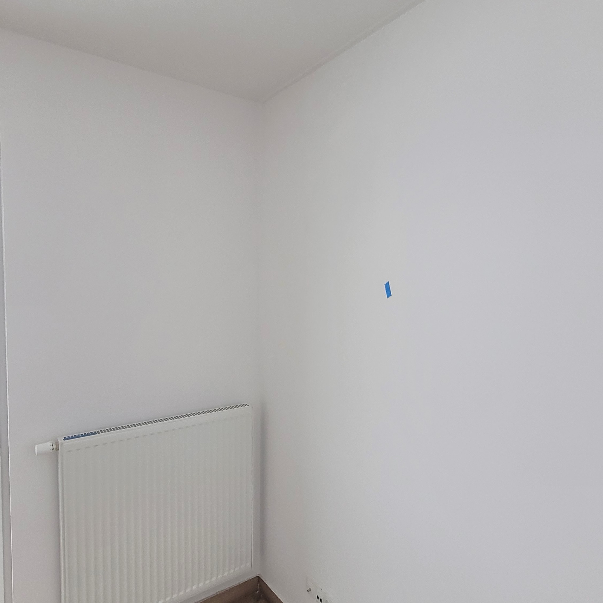 Biała ściana w rogu pokoju z grzejnikiem i małym kawałkiem niebieskiej taśmy. Minimalistyczne wnętrze, świeżo pomalowane ściany, widoczne gniazdka elektryczne przy podłodze.