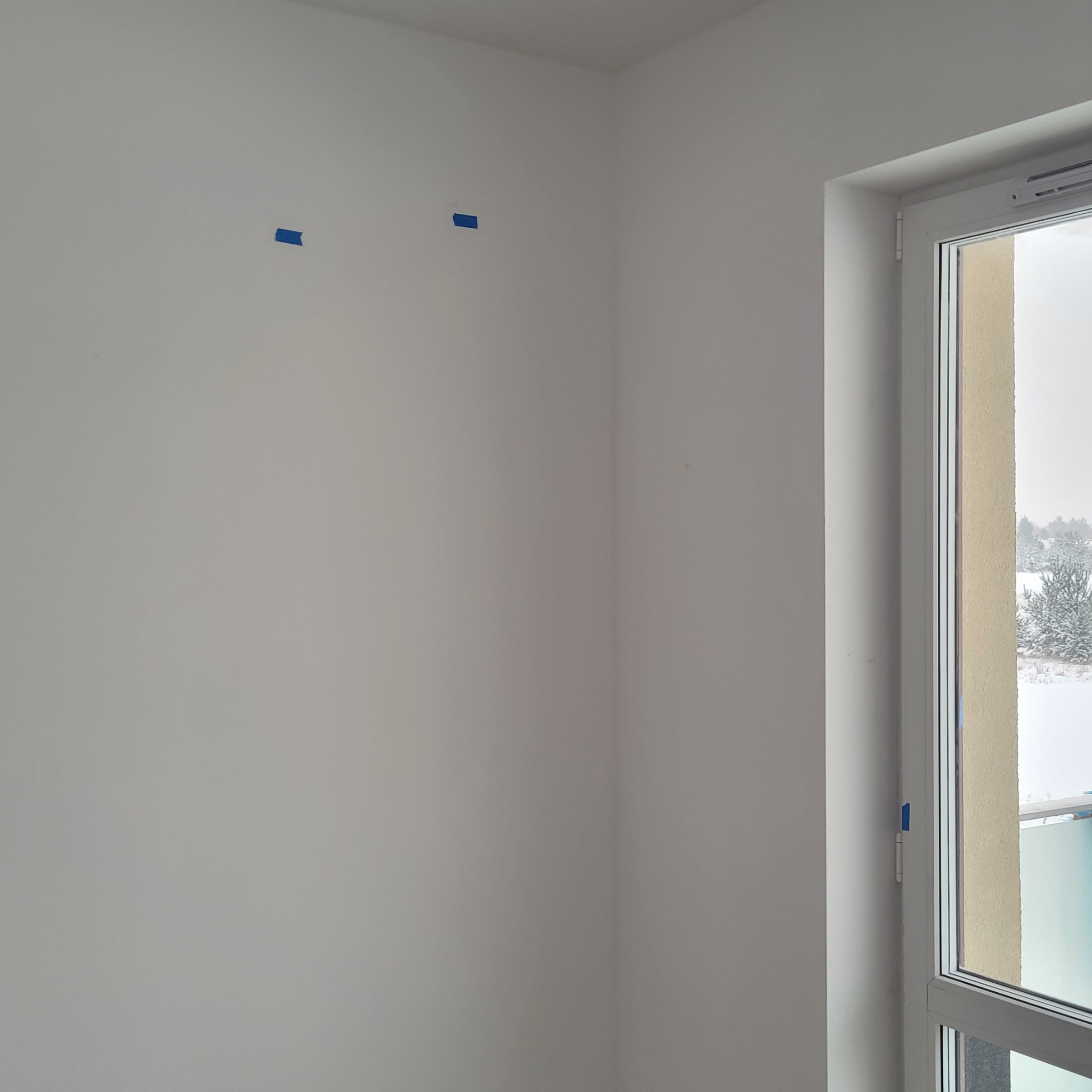 Biała ściana z niebieską taśmą malarską, okno z widokiem na zaśnieżone drzewa. Przygotowanie do malowania ścian w nowym mieszkaniu.