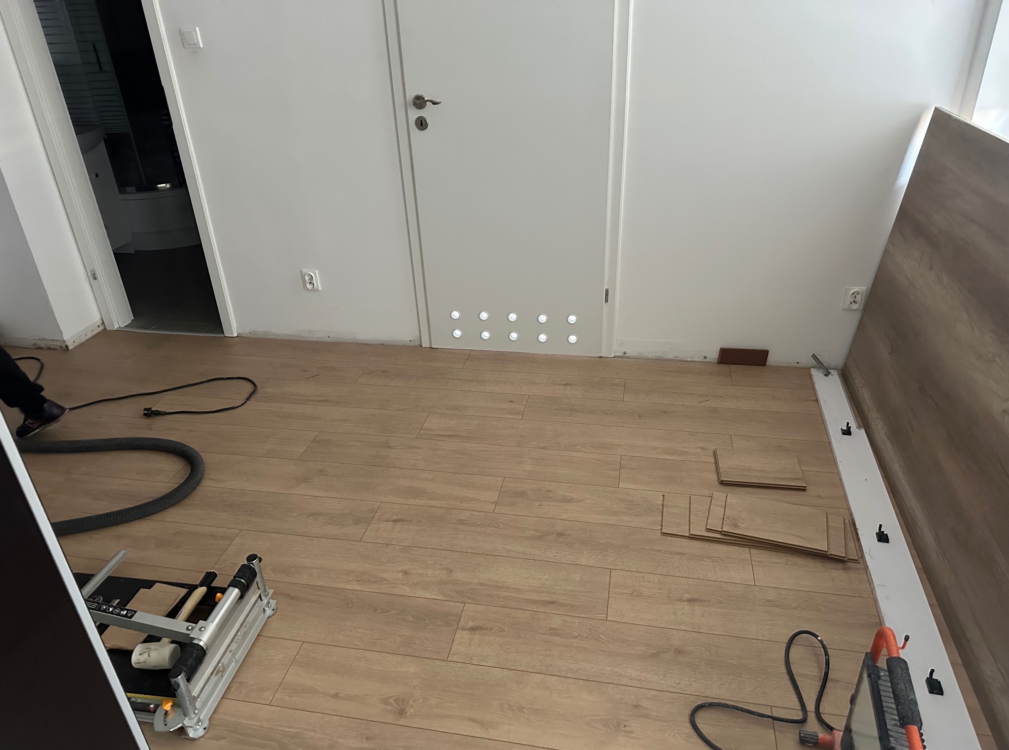 Układanie paneli podłogowych w pokoju, widoczne narzędzia pracy, częściowo ułożone panele i odkurzacz przemysłowy. W tle białe ściany i drzwi z oświetleniem podłogowym.