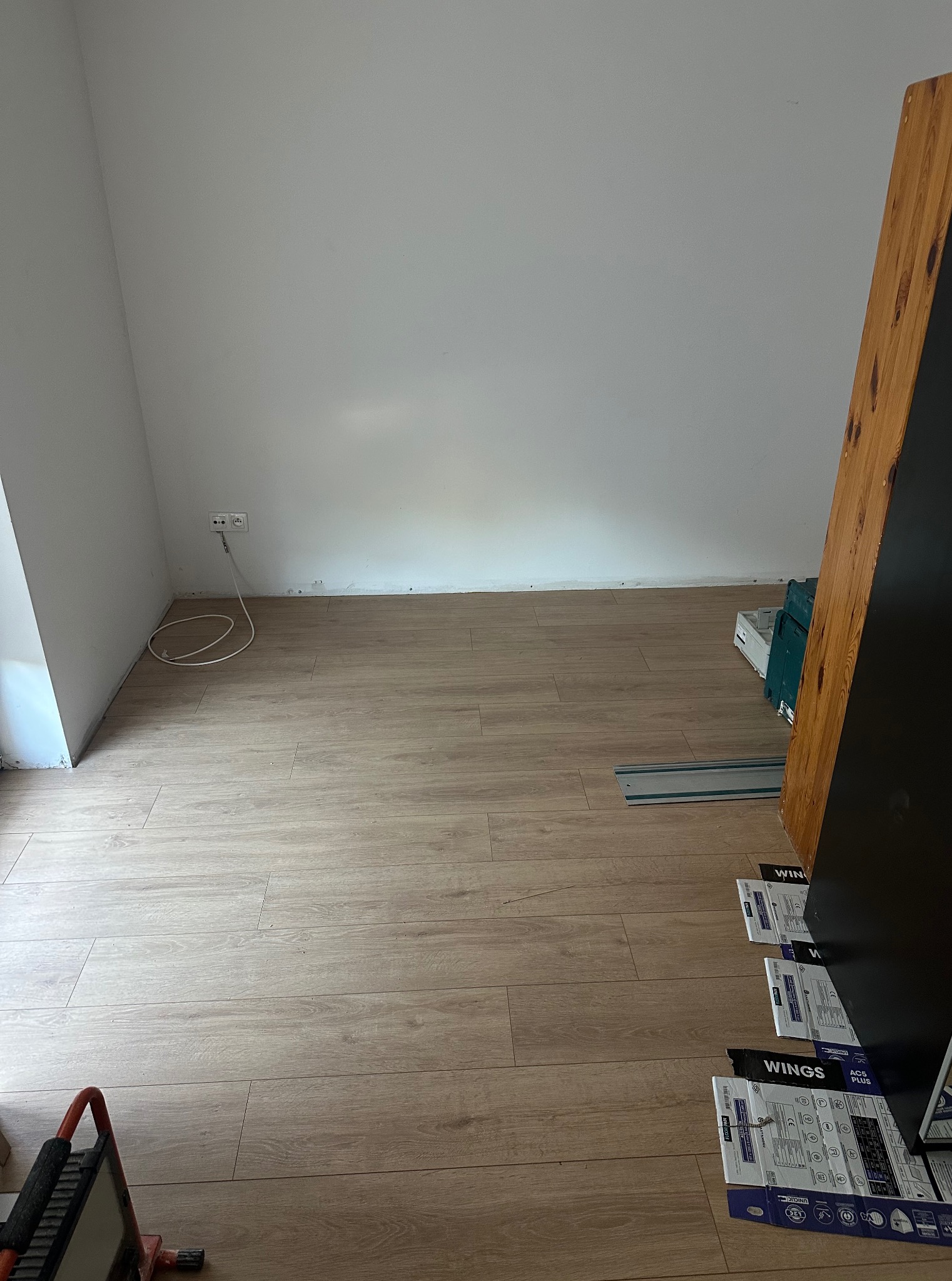 Beżowe panele podłogowe ułożone w pokoju, widoczne narzędzia i opakowania po panelach. Ściany białe, gniazdko elektryczne. Częściowo widoczna szafa.