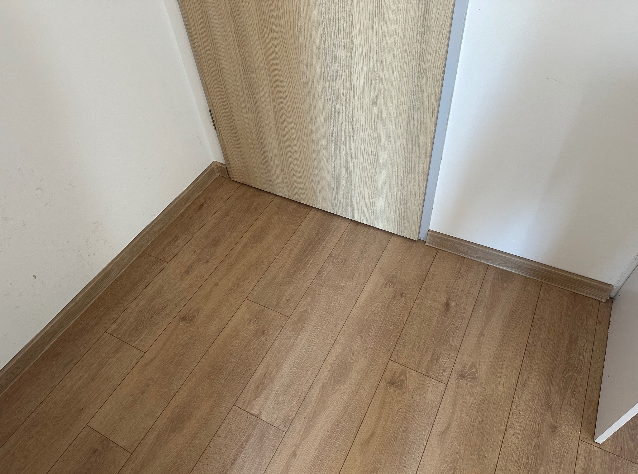 Jasne panele podłogowe ułożone wzdłuż ściany i drzwi z drewnopodobnym wykończeniem, widoczne listwy przypodłogowe.