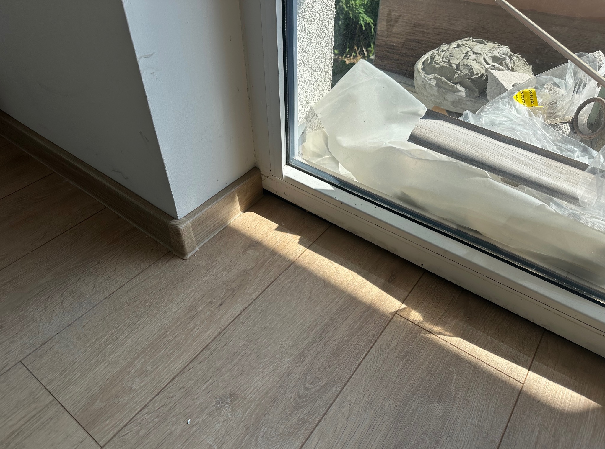 Jasne panele podłogowe z drewnopodobnym wykończeniem ułożone przy białej ścianie i oknie, z widoczną listwą przypodłogową i fragmentem materiałów budowlanych.