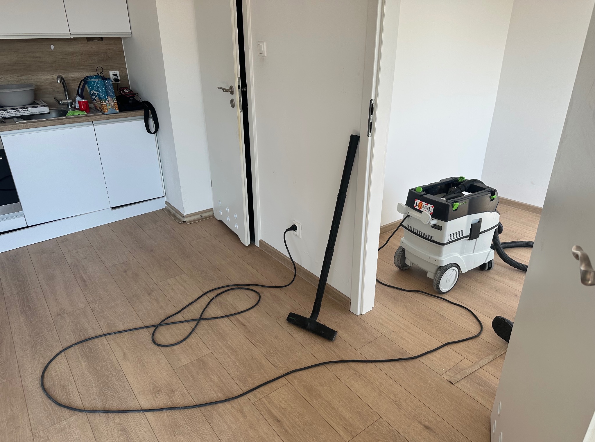 Parkiet po ułożeniu, odkurzacz przemysłowy gotowy do sprzątania. Widoczne panele podłogowe i fragment kuchni. Minimalistyczne wnętrze w trakcie wykończenia.