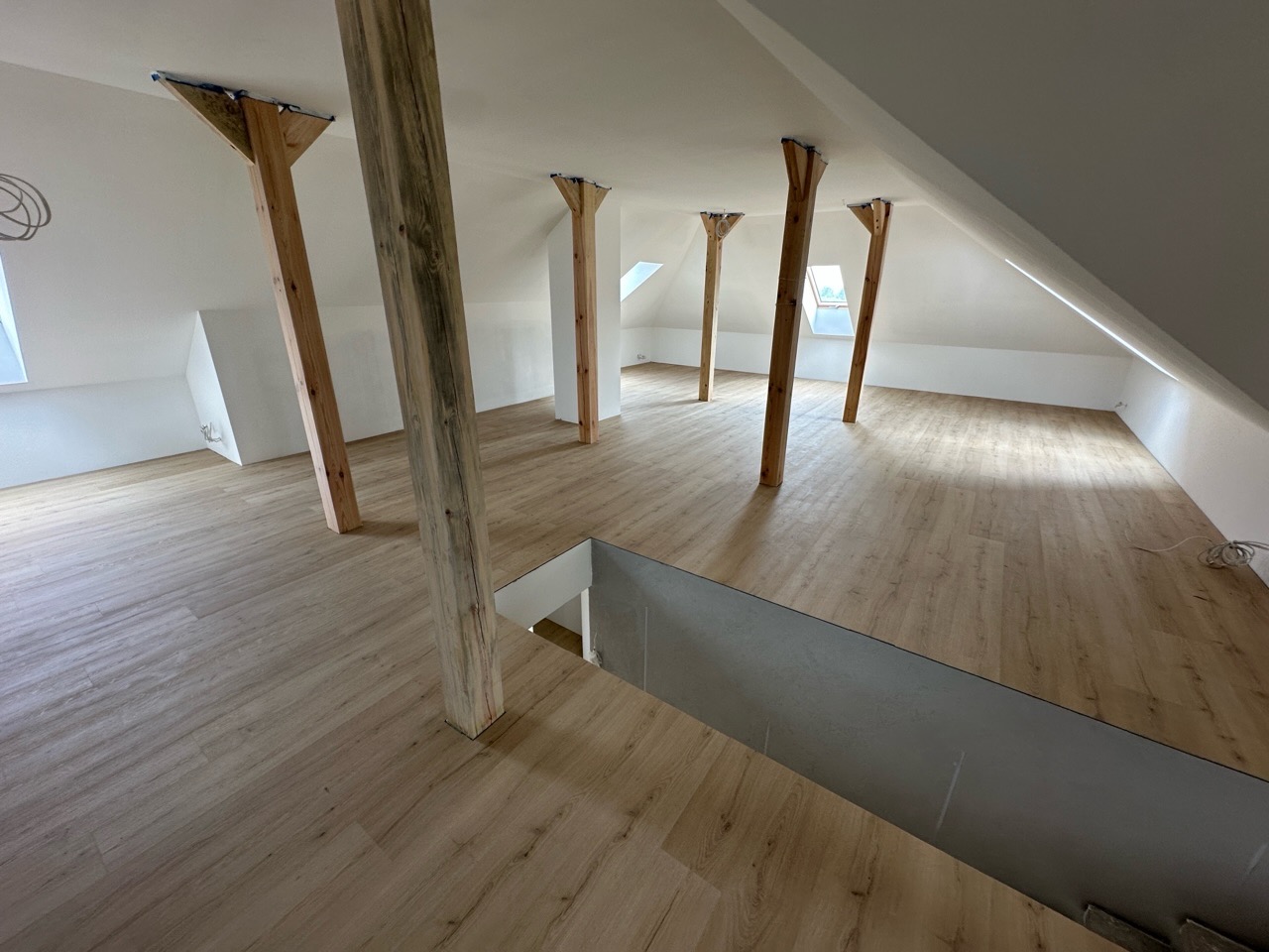 Jasne panele podłogowe na poddaszu z drewnianymi belkami konstrukcyjnymi i oknami dachowymi. Widok na wykończone wnętrze z perspektywy schodów.