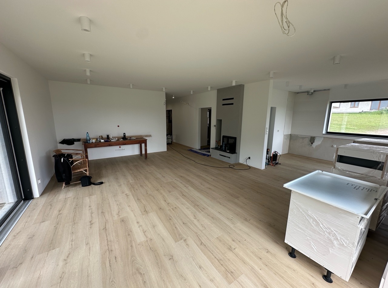 Jasne panele podłogowe w przestronnym salonie z minimalistycznym kominkiem i oknem. Stolik z narzędziami w tle, elementy wyposażenia zabezpieczone folią.