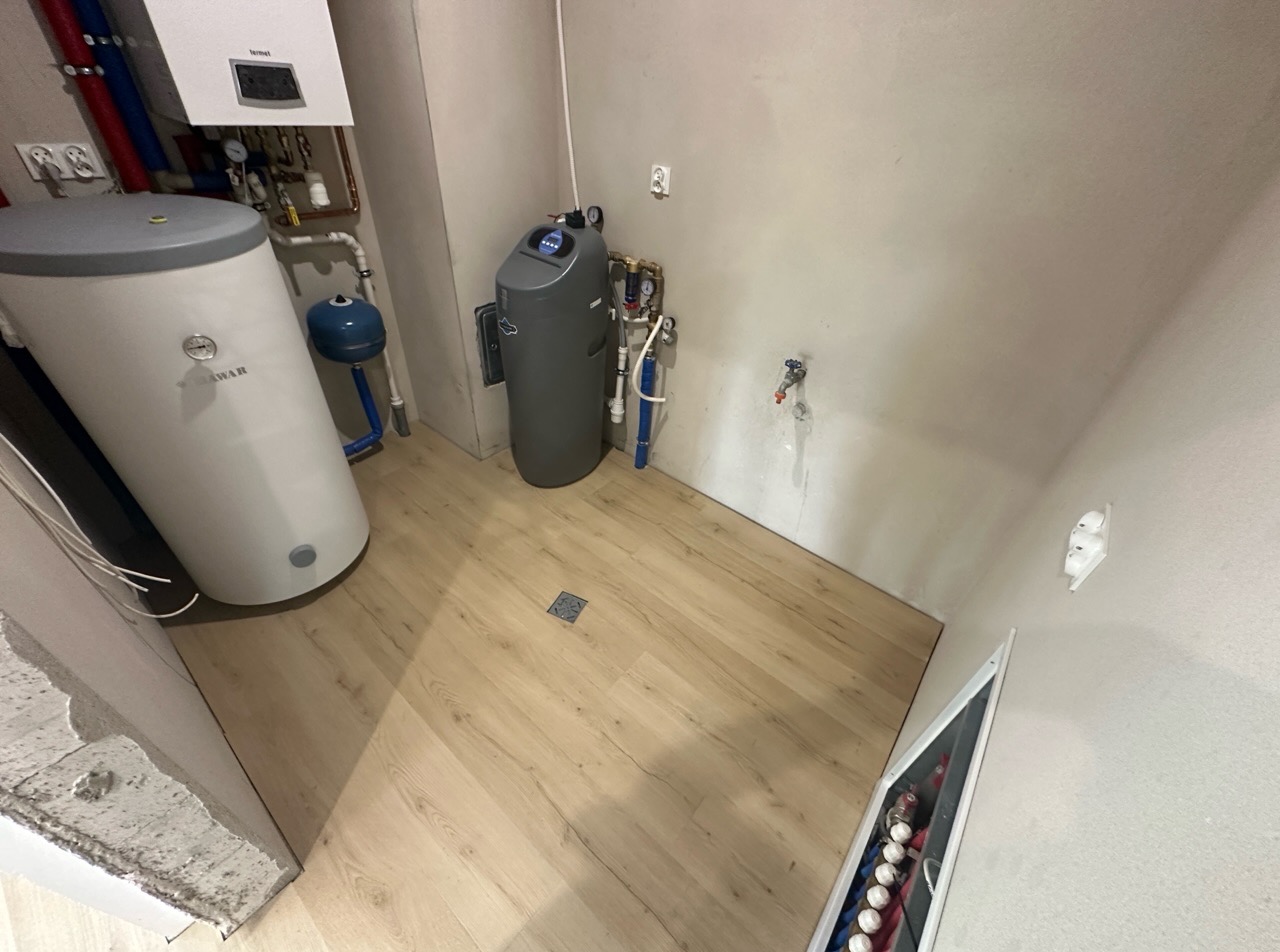 Jasne panele podłogowe w pomieszczeniu technicznym z bojlerem i innymi instalacjami, widoczne przyłącza wodne i elektryczne.