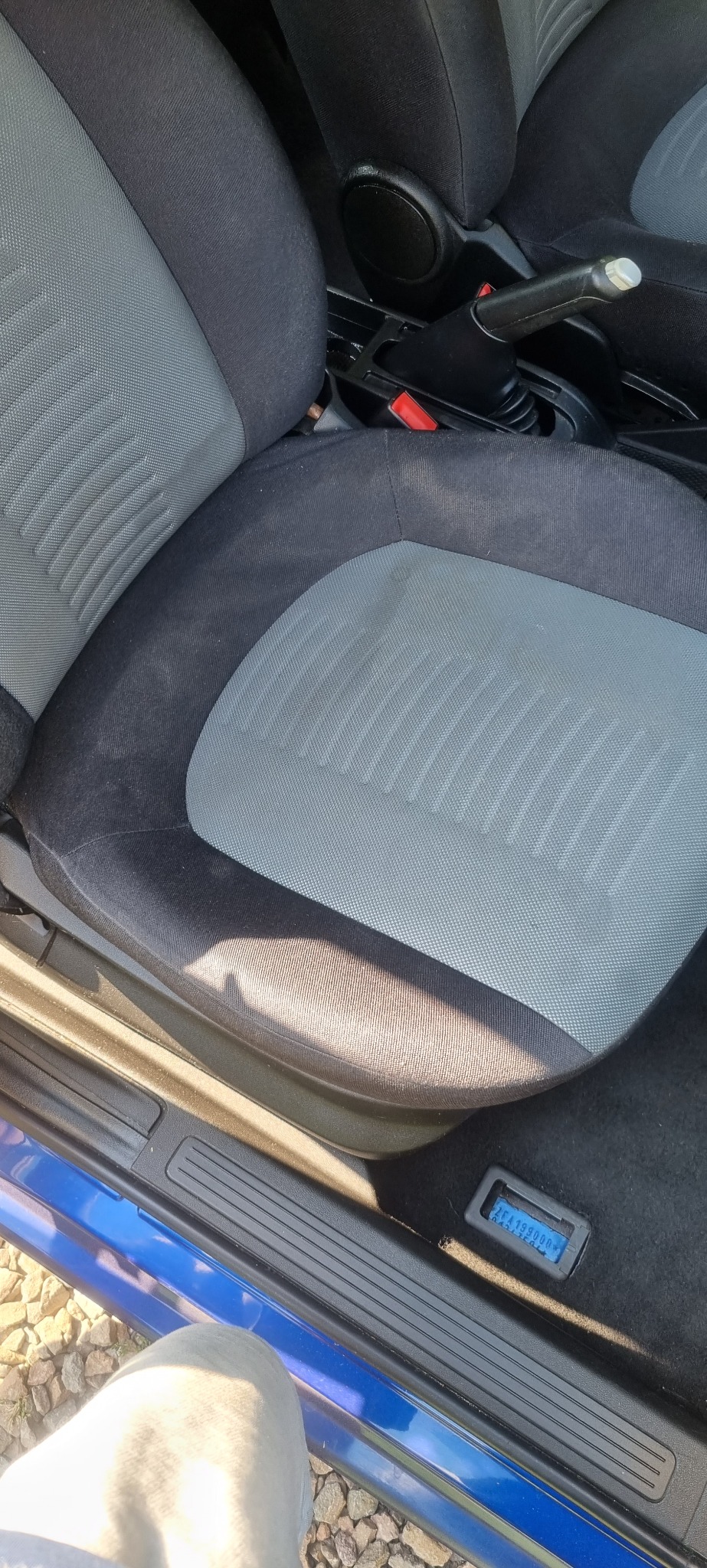 Szare, tekstylne siedzenie samochodowe z widocznymi plamami i zabrudzeniami, sugerujące potrzebę wyczyszczenia tapicerki.