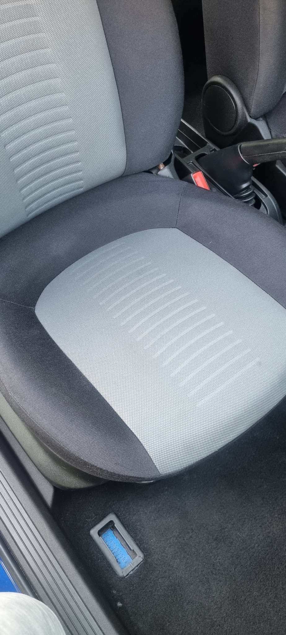 Szare, tekstylne siedzenie samochodowe z widoczną fakturą i wzorem w paski, z elementami czarnej tapicerki. Widoczny fragment podłogi i konsoli środkowej.
