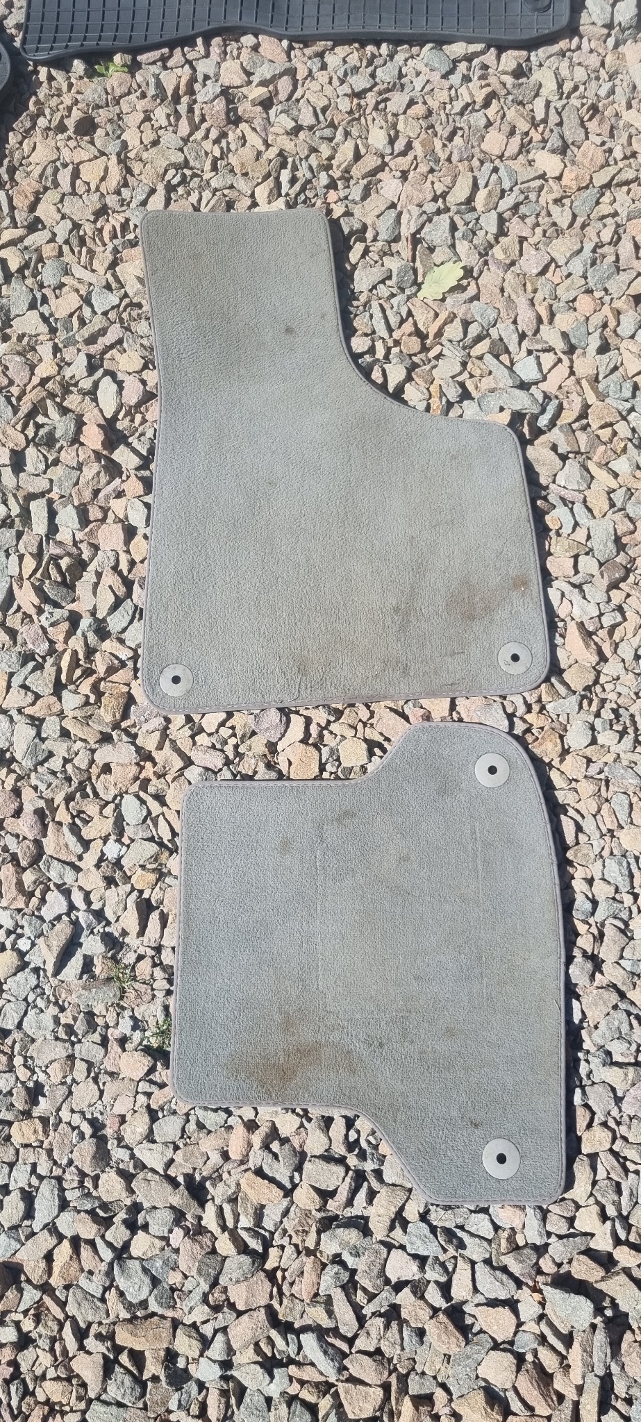 Dwa brudne, szare dywaniki samochodowe z mocowaniami na żwirowym podłożu. Widoczne plamy i zabrudzenia na powierzchni materiału.