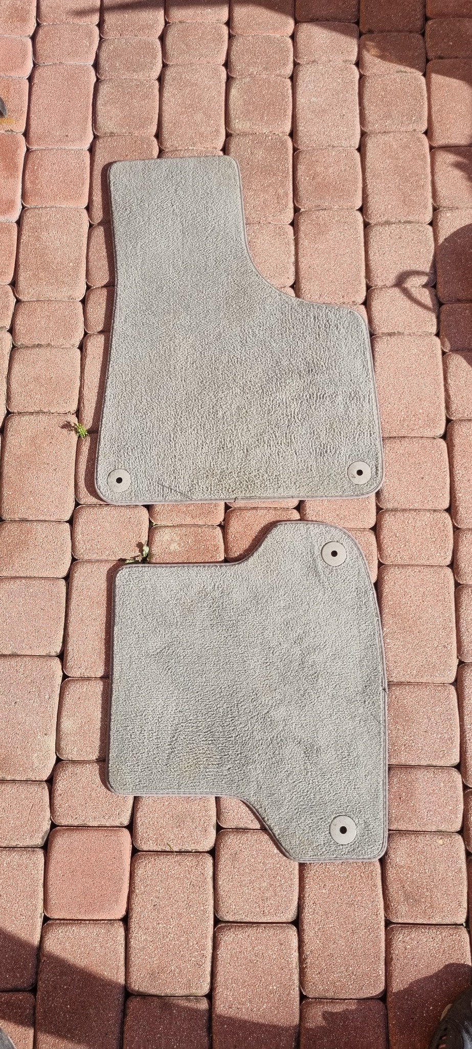 Dwa wyprane, jasnoszare dywaniki samochodowe z mocowaniami, leżące na czerwonej kostce brukowej w słoneczny dzień. Widoczne drobne ślady użytkowania.