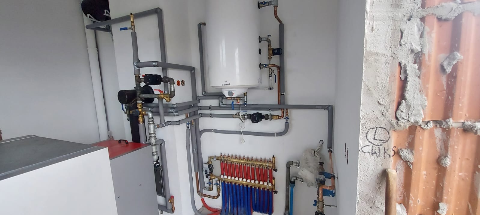 Złożona instalacja hydrauliczna z rozdzielaczem, rurami w izolacji i bojlerem w białym pomieszczeniu z surową ścianą z cegły. Elementy miedziane i zawory.