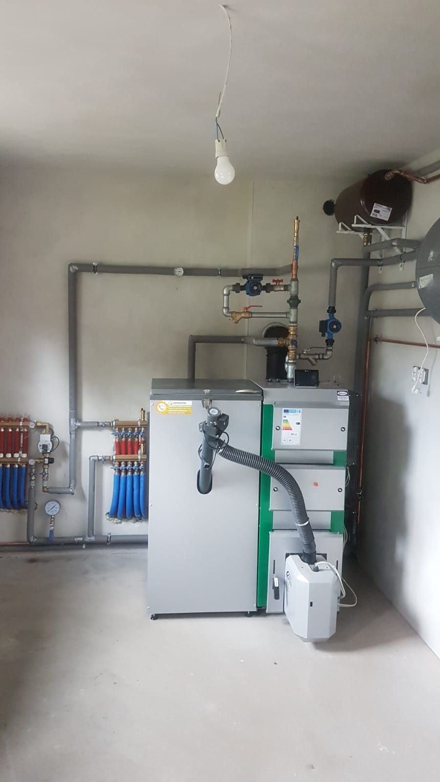Zmontowana instalacja grzewcza z kotłem, zasobnikiem i rozdzielaczem, rury rozprowadzone po ścianach, surowy stan wykończenia pomieszczenia, żarówka na kablu.