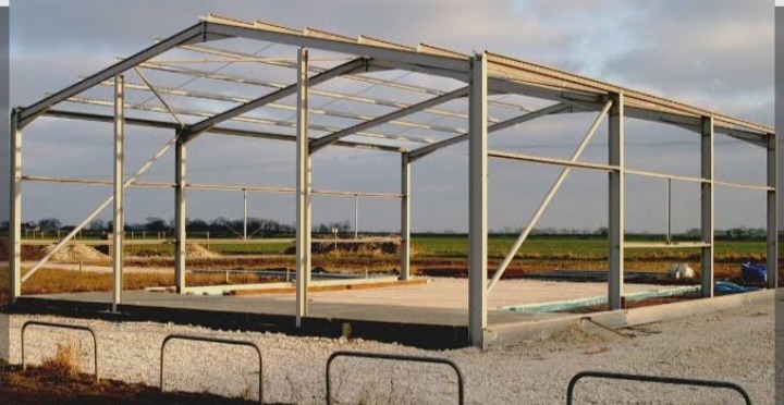 Stalowy szkielet hali na betonowej płycie fundamentowej, widok z zewnątrz na tle zielonych pól uprawnych, pochmurne niebo.