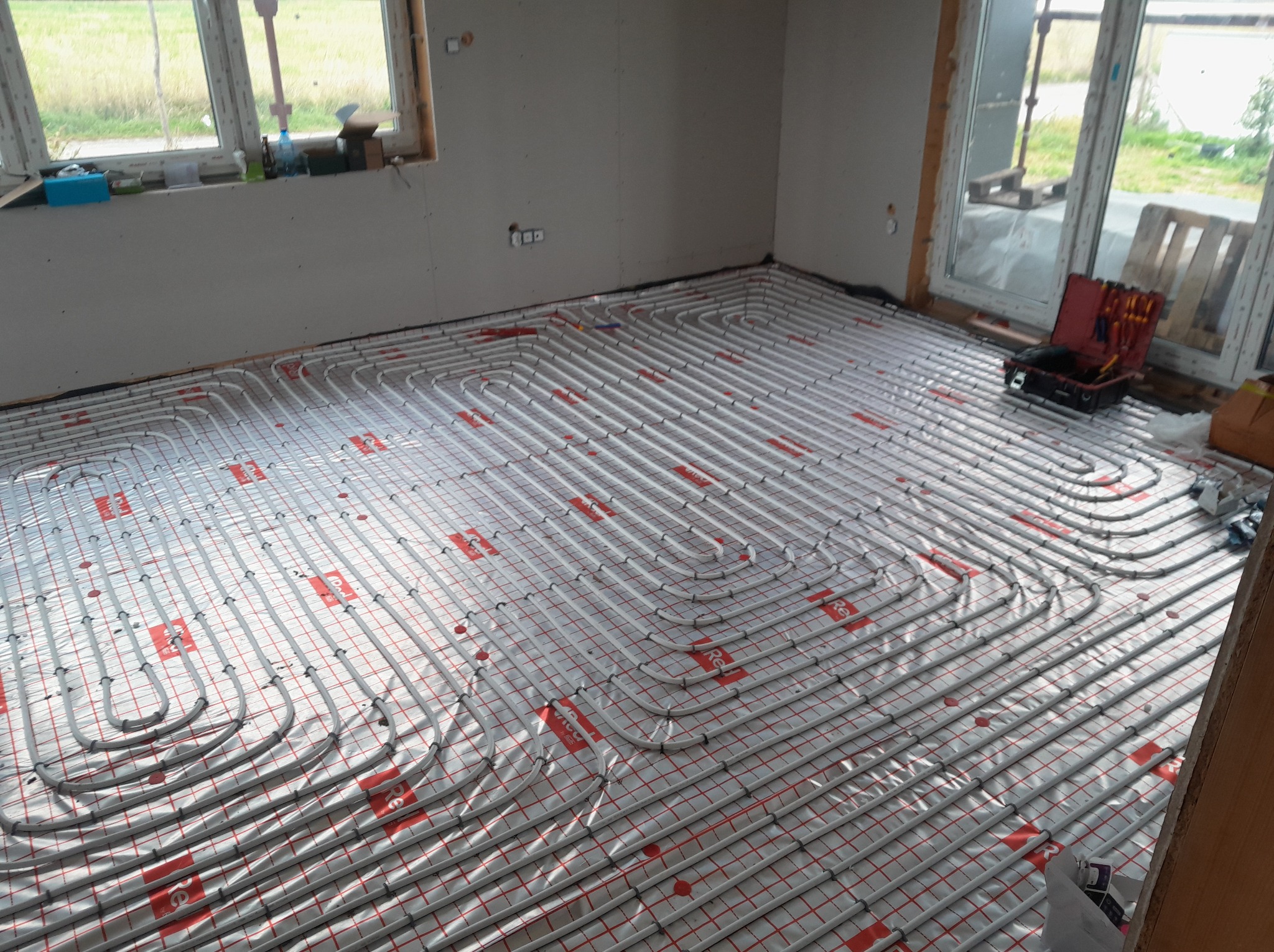 Rozłożone rury ogrzewania podłogowego na folii z czerwoną siatką w surowym wnętrzu, widoczna skrzynka z narzędziami i okno. Etap prac instalacyjnych.