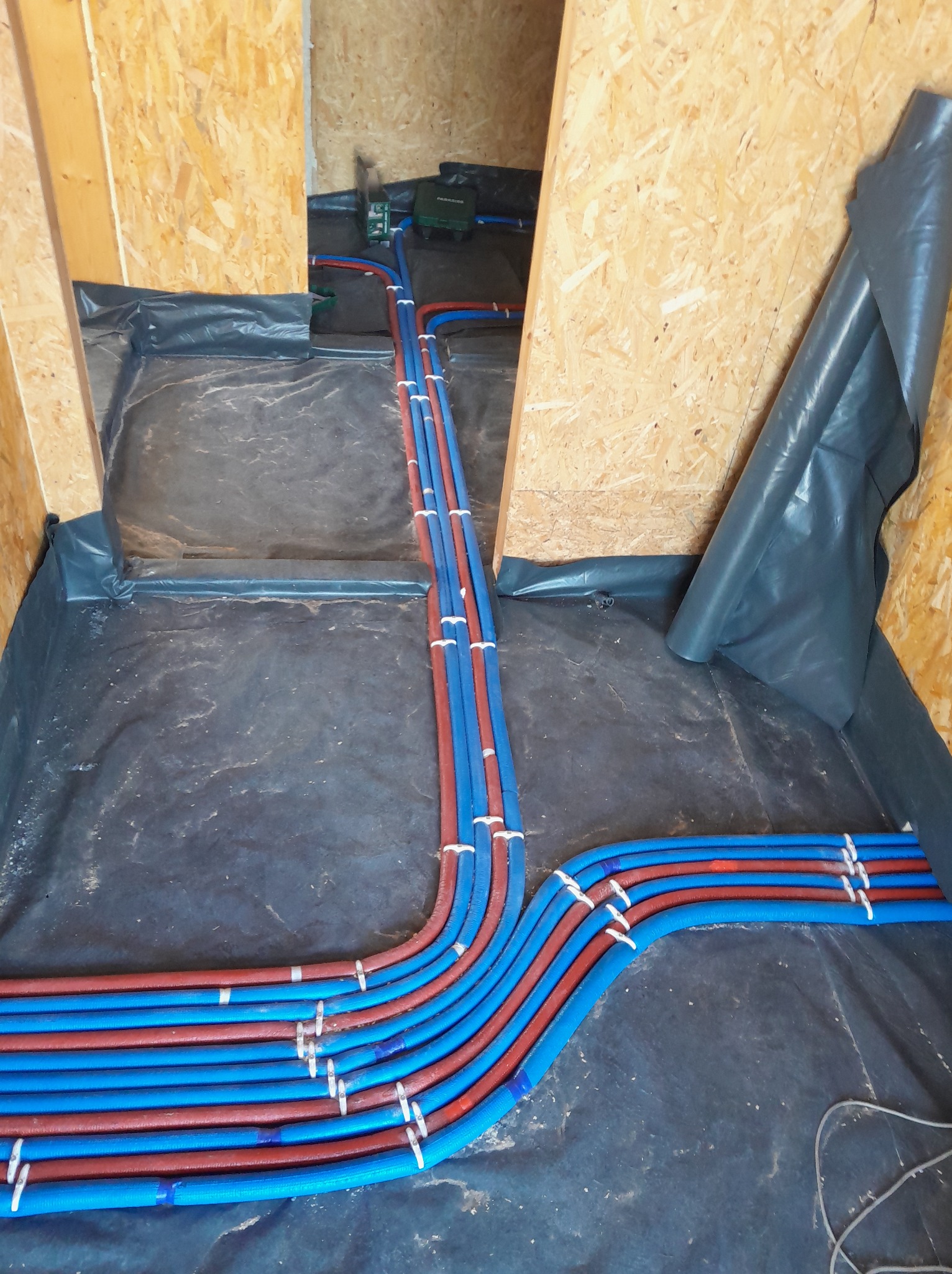 Instalacja ogrzewania podłogowego: rury w kolorze niebieskim i czerwonym ułożone na czarnej folii izolacyjnej w surowym wnętrzu z płyt OSB.