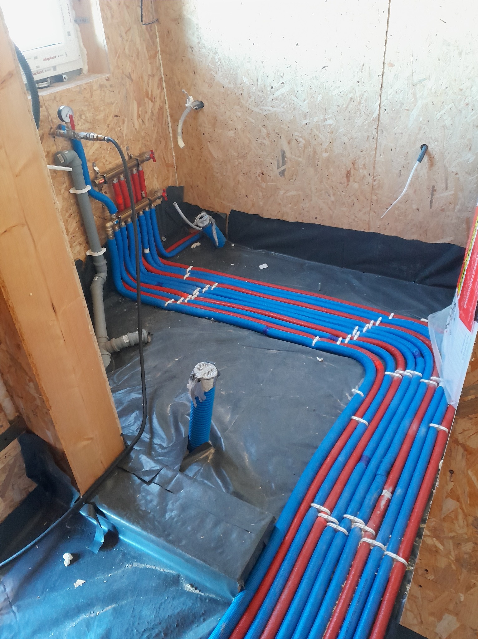 Instalacja ogrzewania podłogowego w surowym wnętrzu: rozłożone rury grzewcze w kolorach niebieskim i czerwonym na czarnej folii izolacyjnej, przed wylaniem posadzki.