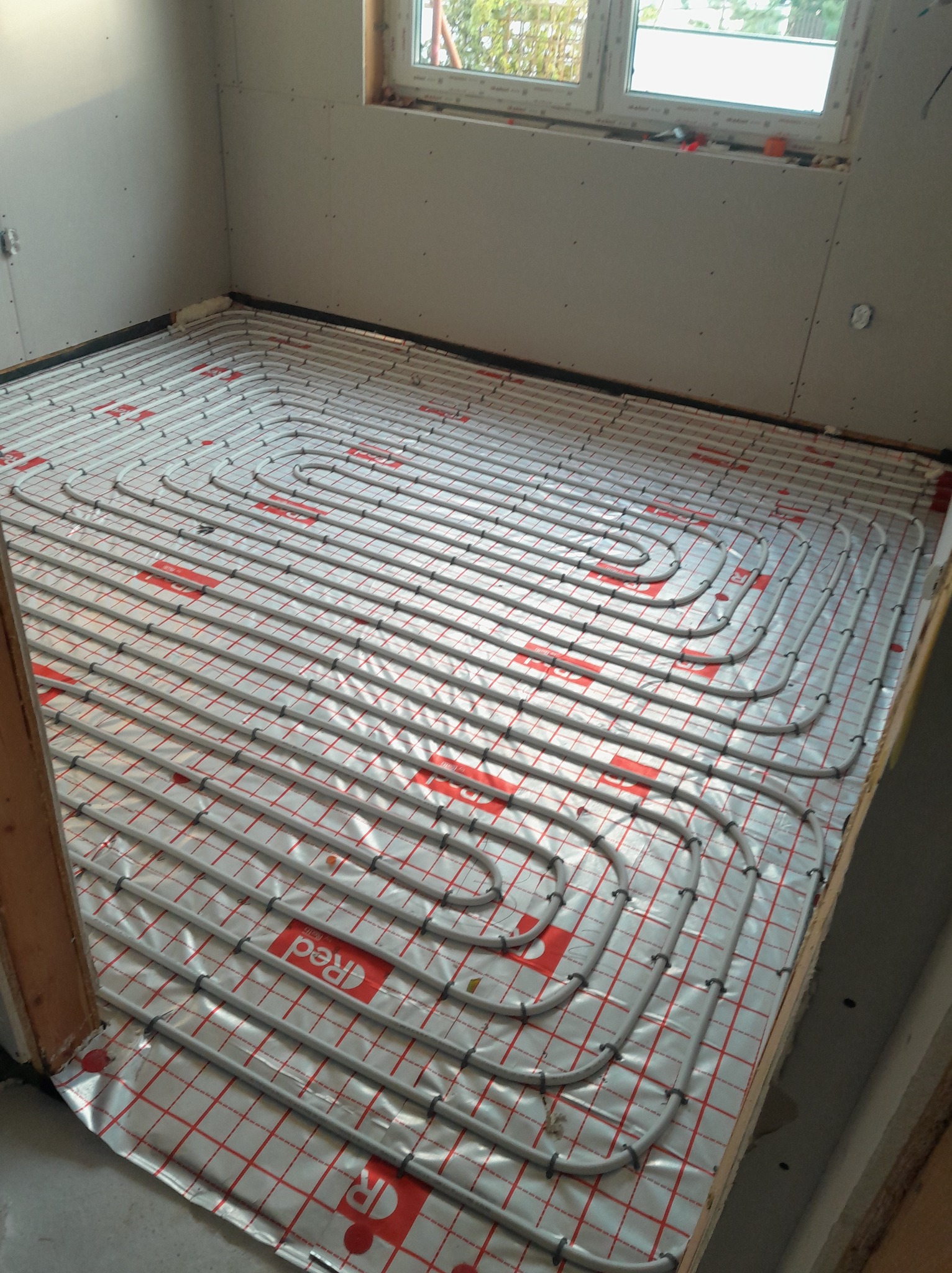 Instalacja ogrzewania podłogowego w pomieszczeniu z oknem. Równomiernie ułożone rury na folii z czerwoną kratką, gotowe do zalania posadzką. Widok z góry.