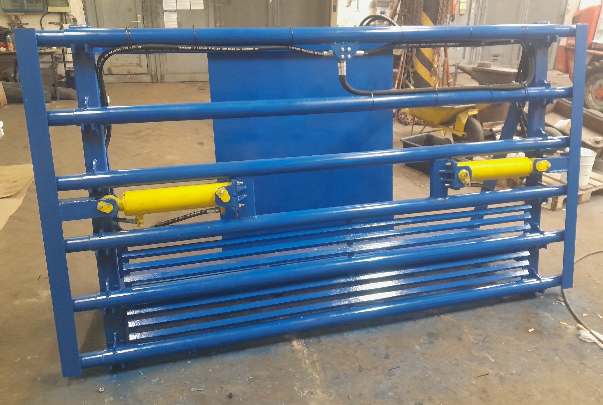 Niebieska, spawana konstrukcja stalowa z żółtymi siłownikami hydraulicznymi, widoczna w warsztacie. Element maszyny rolniczej lub przemysłowej.