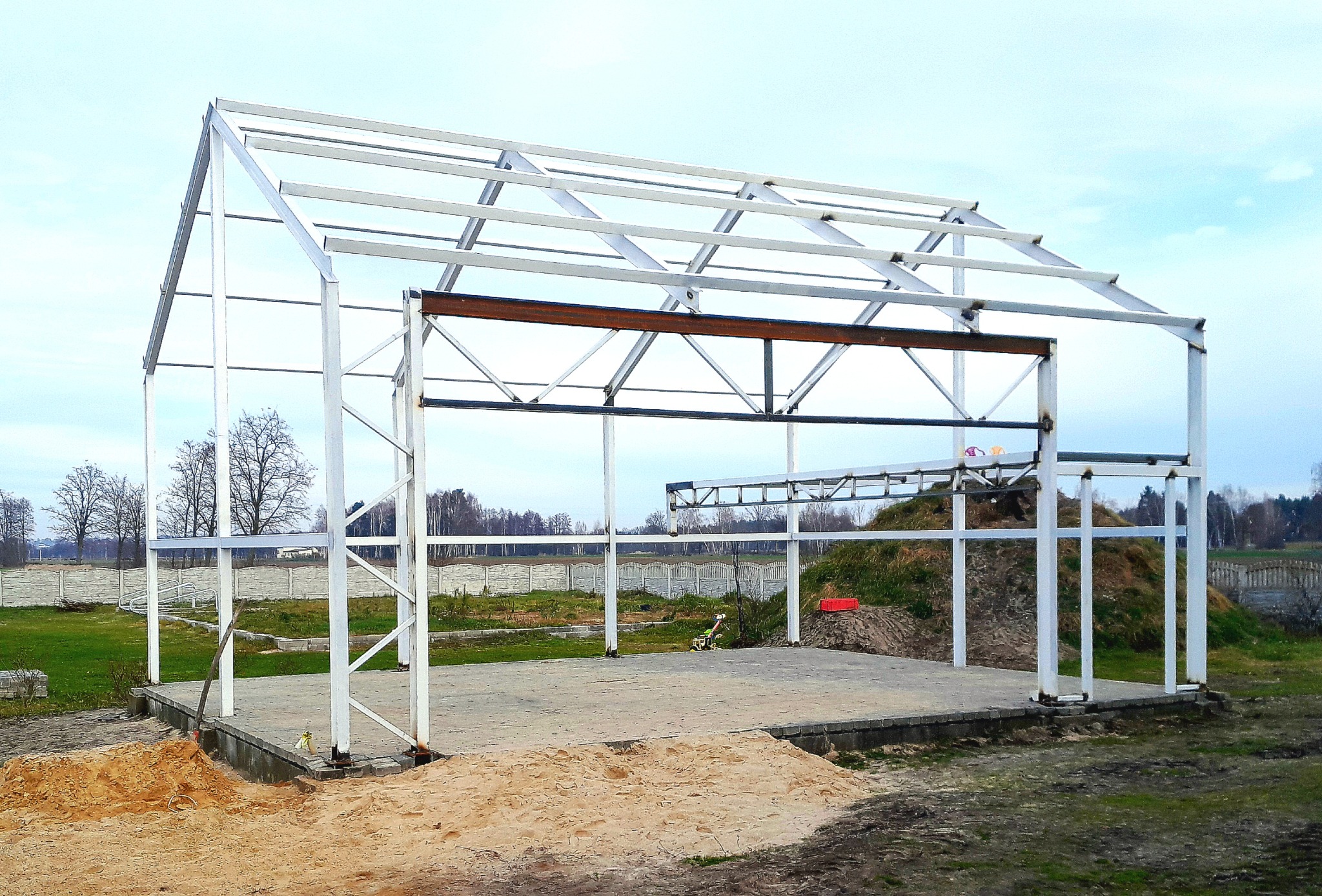 Stalowa konstrukcja hali w trakcie budowy, widok z zewnątrz na betonową podstawę i szkielet z białych profili, w tle drzewa i ogrodzenie.