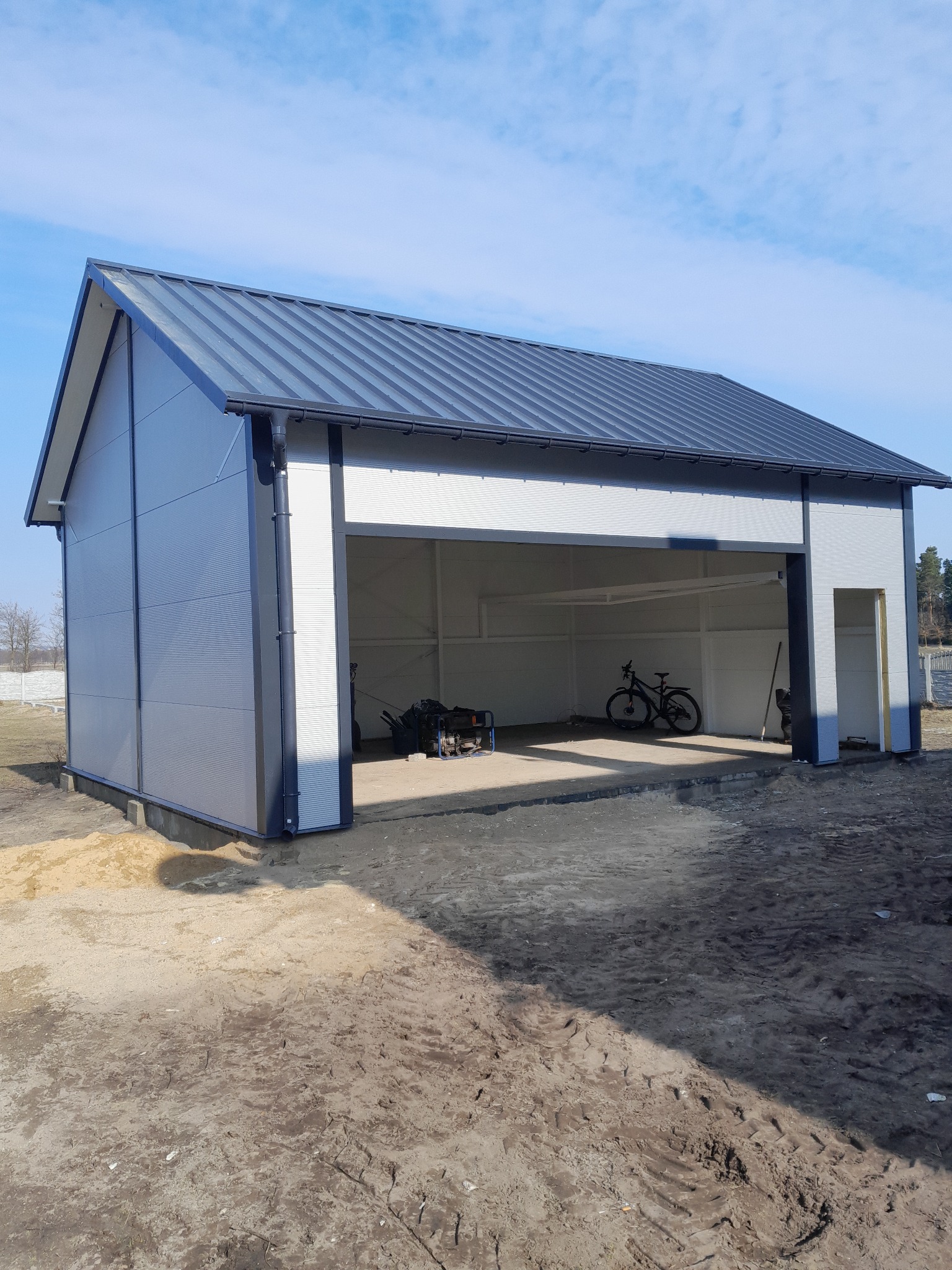 Nowoczesna hala z płyt warstwowych w odcieniach szarości, z otwartą bramą i widocznym wnętrzem. Na zewnątrz teren przygotowany pod dalsze prace.