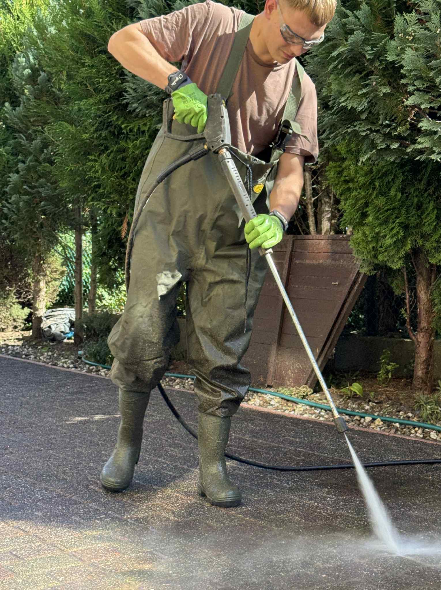 Mężczyzna w wodoszczelnym kombinezonie i okularach ochronnych czyści kostkę brukową myjką ciśnieniową na tle zielonych krzewów i drzew.