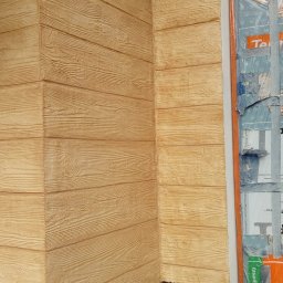 Elewacje Ocieplenia DomówTynki Natryskowe Koszalin, Kołobrzeg SUB 177 Budowlane - Elewacja budynku z fakturą drewna, zabezpieczone okno folią malarską. Widoczny narożnik i fragment instalacji. Ciepłe, naturalne odcienie brązu i beżu.